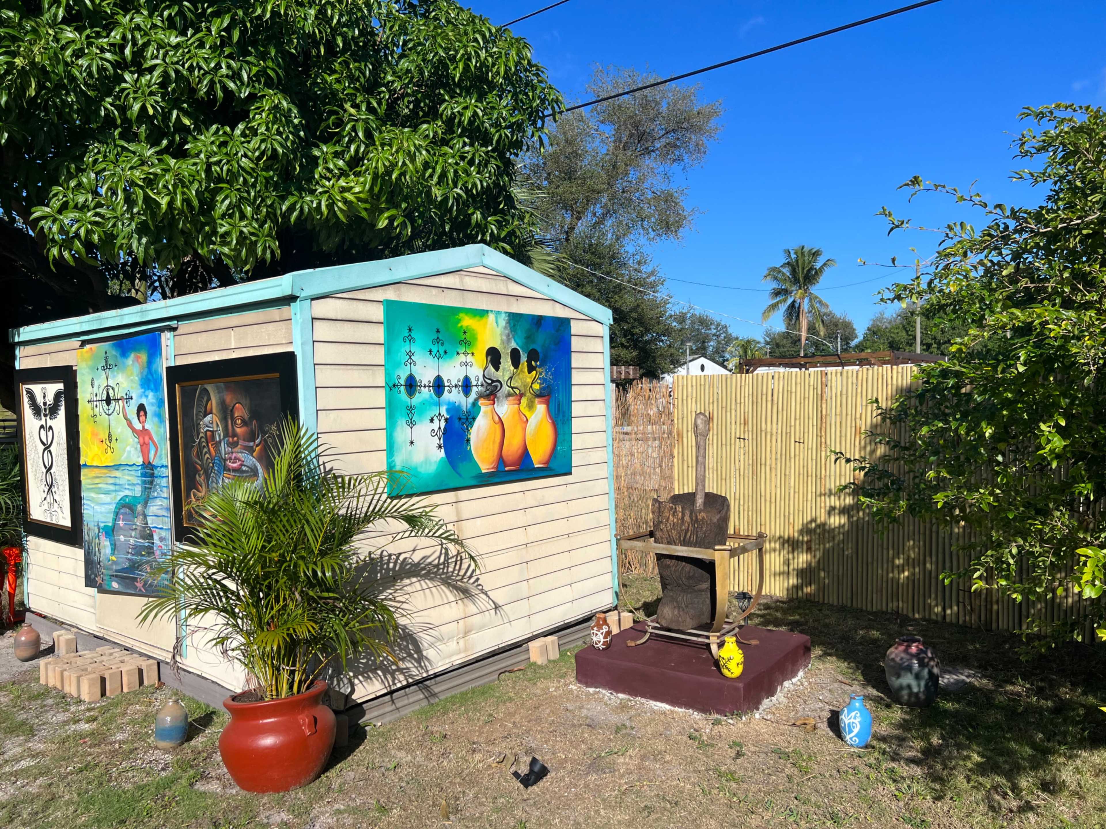 A small wooden shed with colorful artwork on the walls is surrounded by plants and decorative items in a yard.