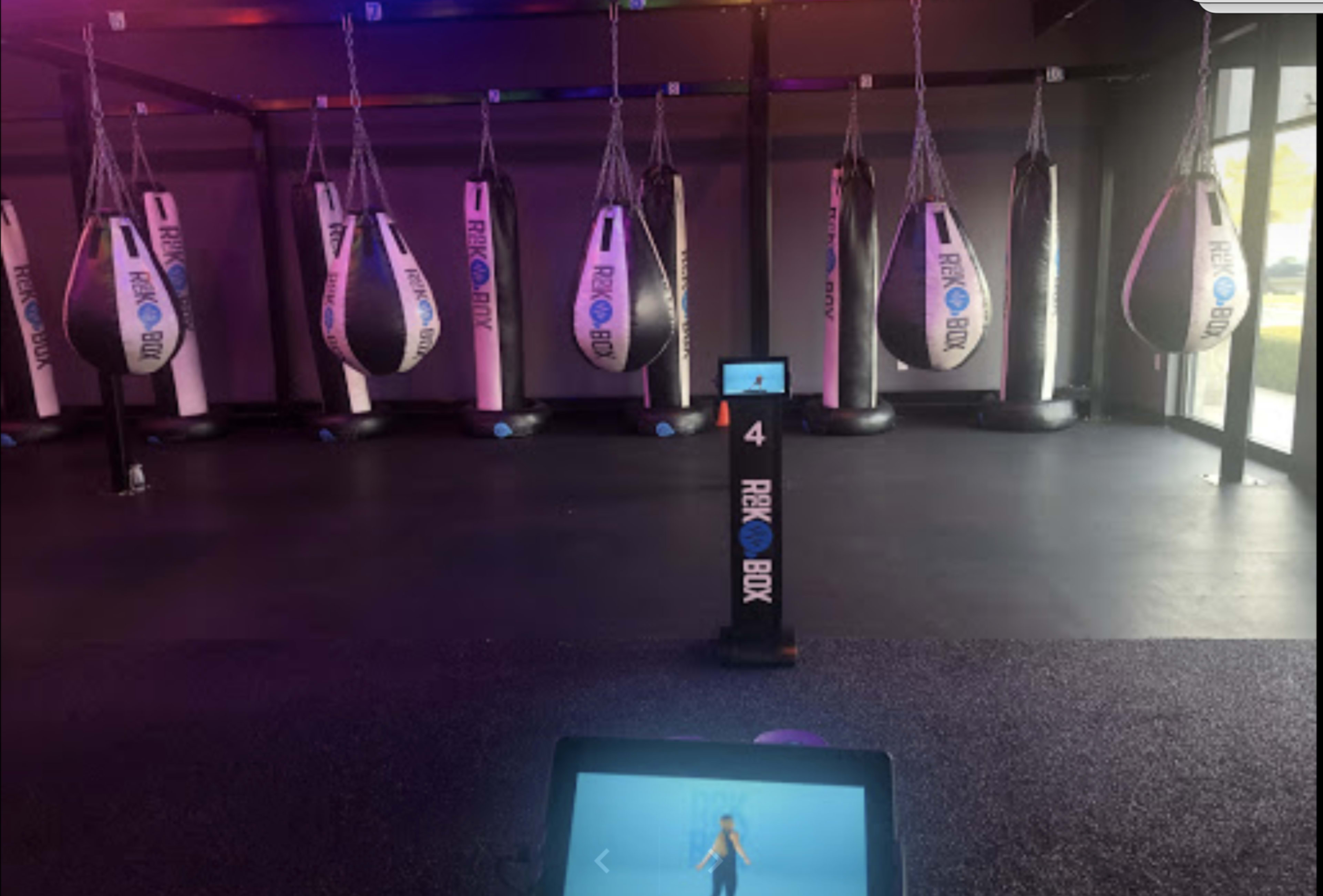 A boxing gym interior featuring hanging heavy bags and a display monitor on a stand.