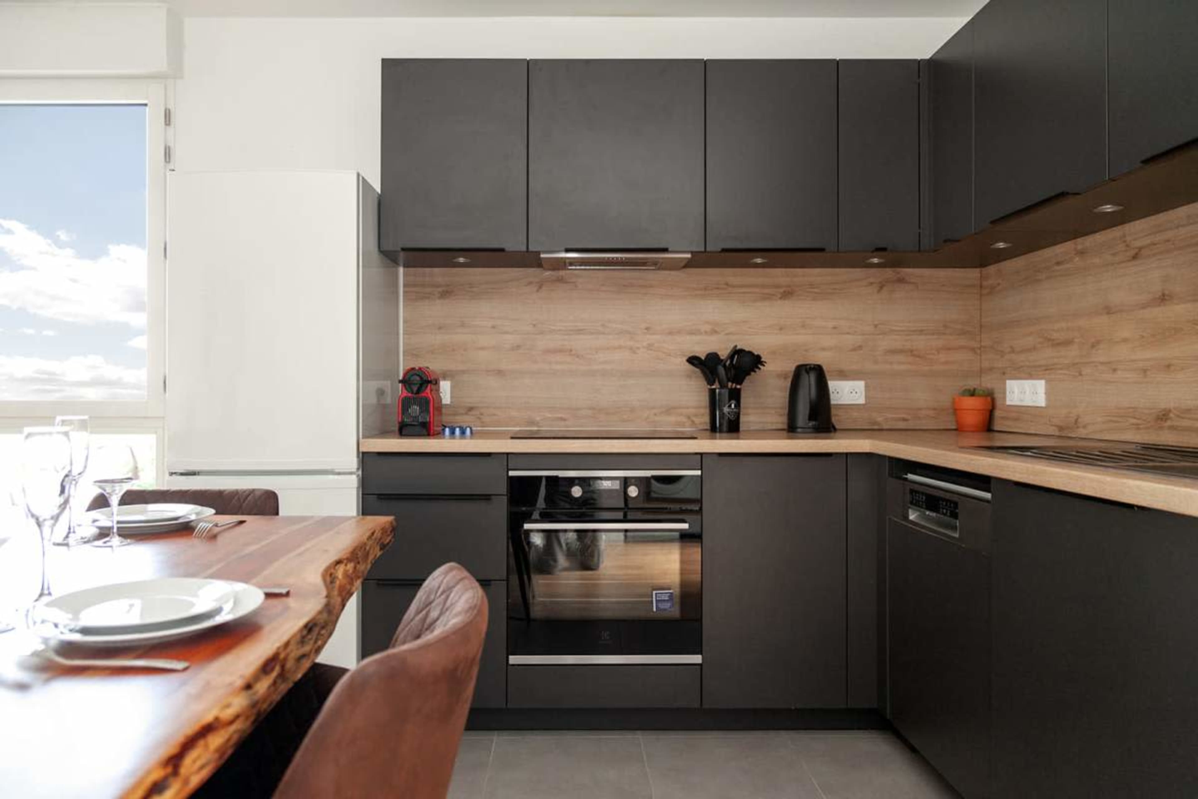 The image shows a modern kitchen with dark cabinetry, a wooden backsplash, and a dining area featuring a wooden table set for a meal.