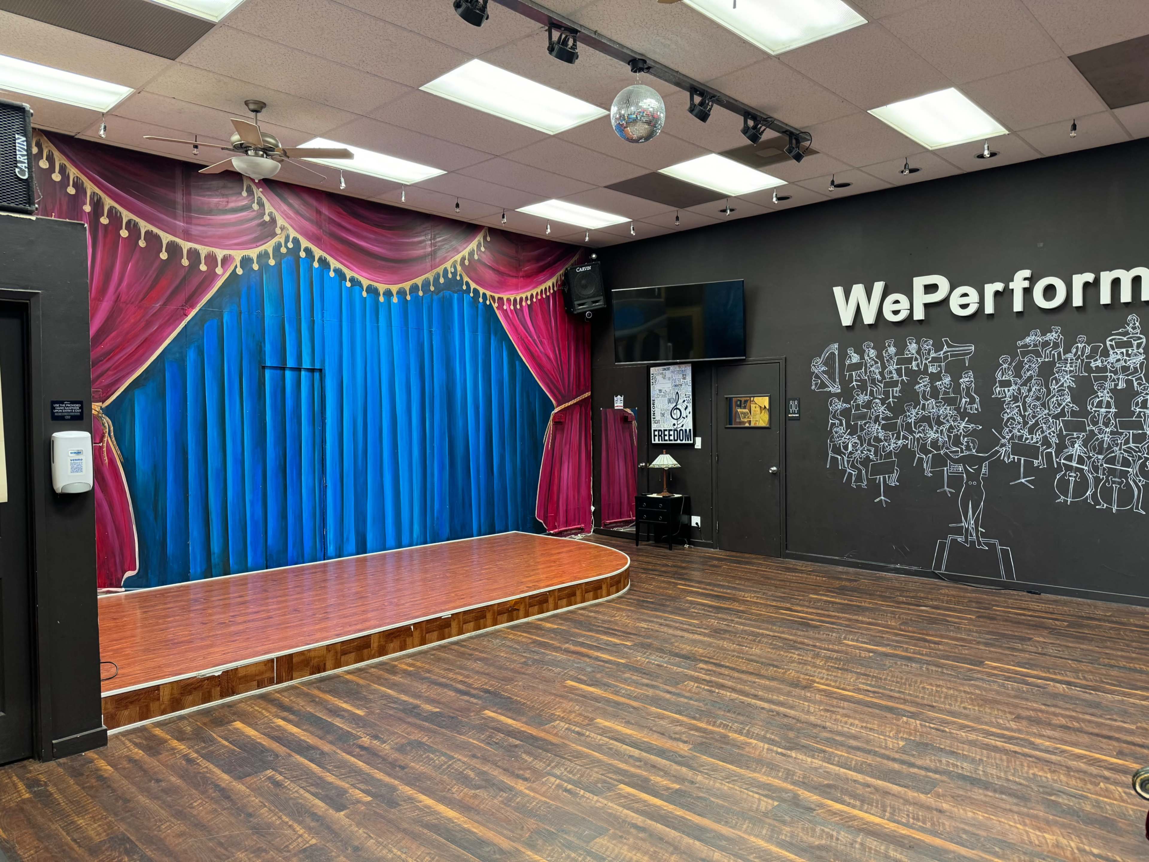 The image shows a stage with a blue curtain backdrop and red velvet drapes, set in a room featuring a wooden floor and a wall adorned with musical illustrations.