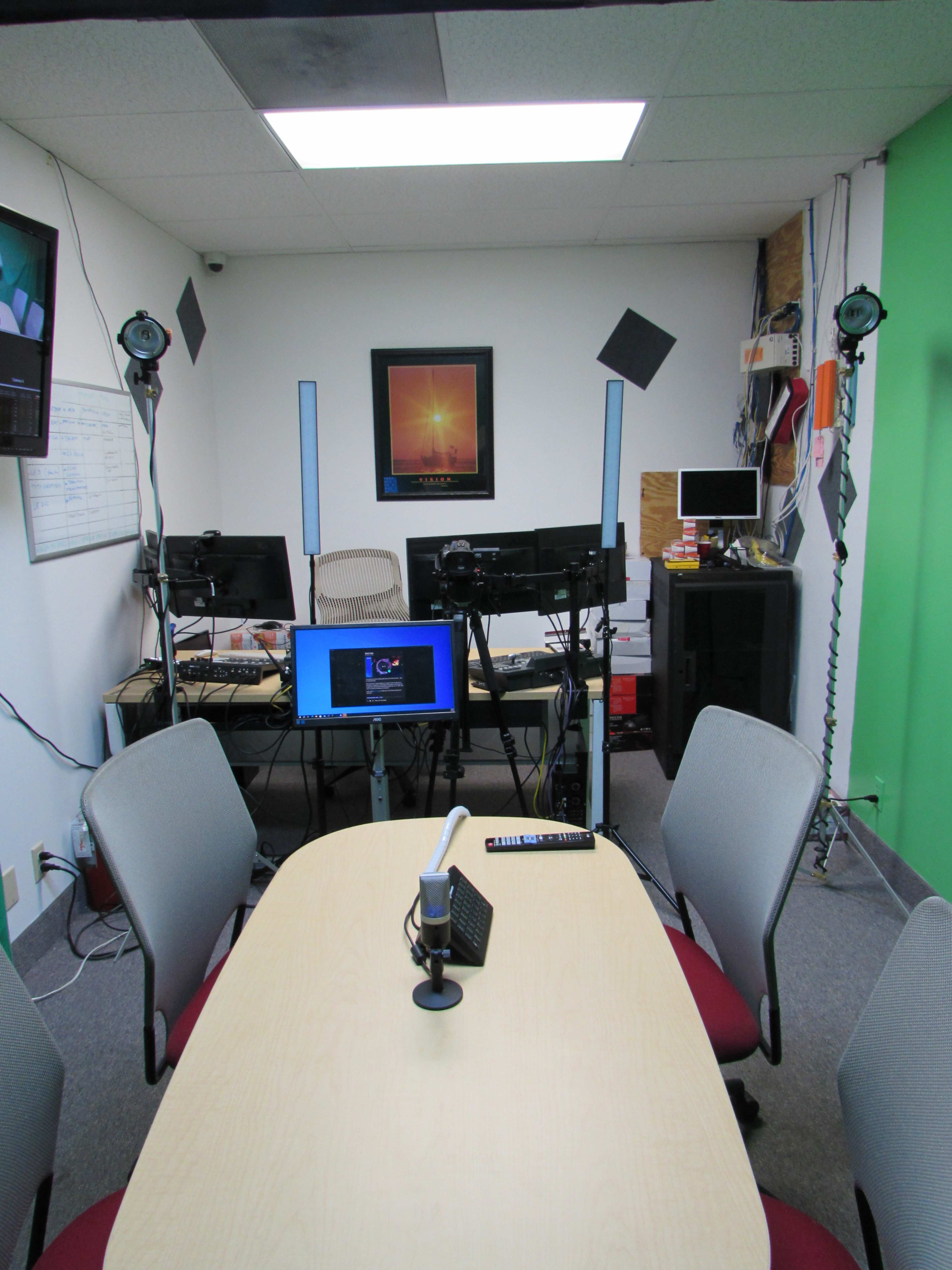 The image shows a small, well-equipped studio with a table, chairs, computers, and lighting setups, along with a green screen on one wall.
