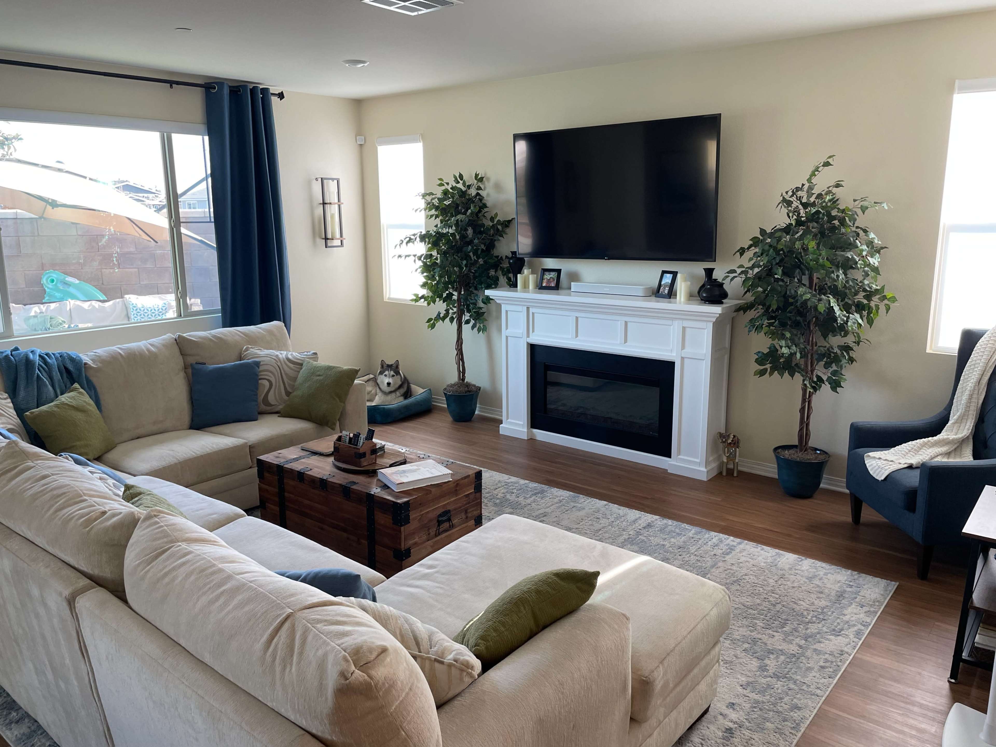 A living room featuring a beige sectional sofa, a wooden coffee table, two potted plants, a wall-mounted TV, and a decorative fireplace.