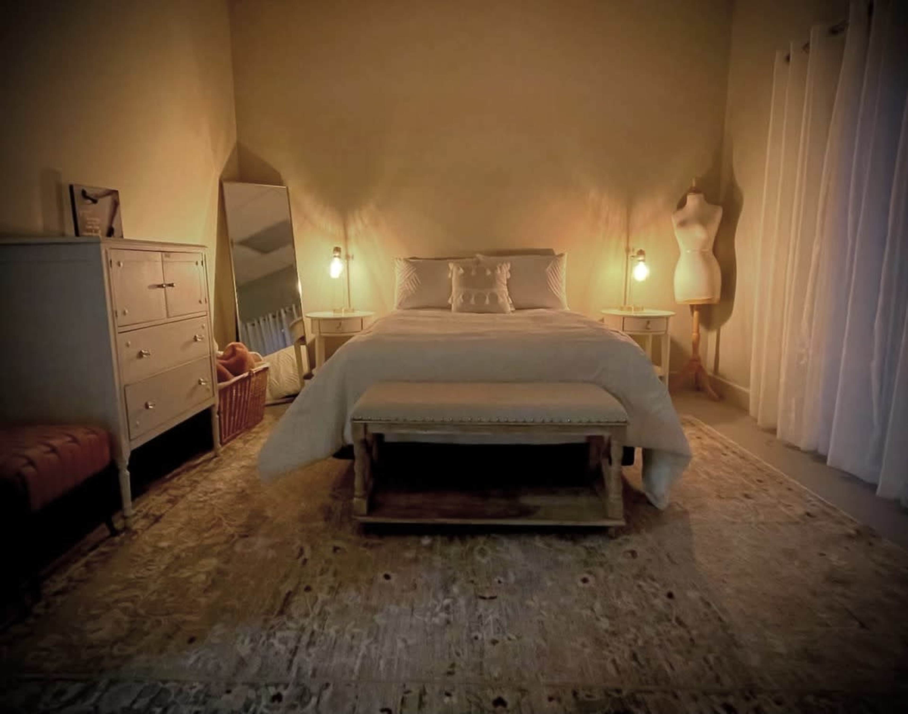 The image shows a softly lit bedroom featuring a bed, a dresser, a floor mirror, and a clothing mannequin beside a window.