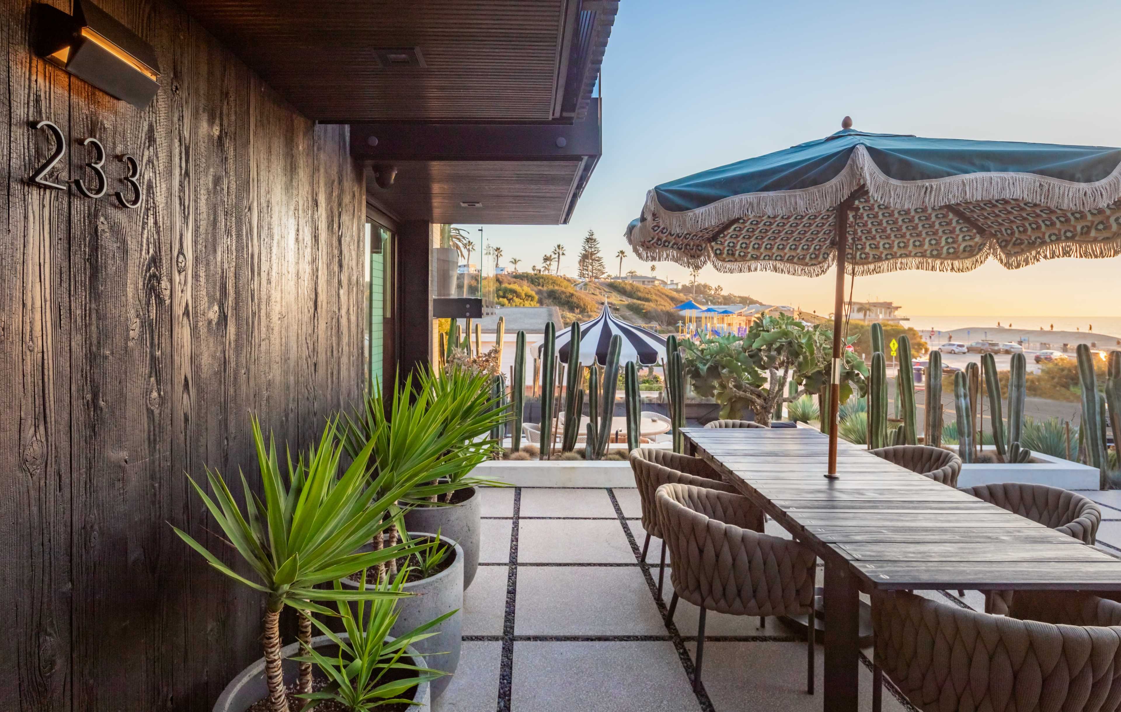 A patio area features a large table under a decorative umbrella, surrounded by potted plants, with a view of the beach in the background.