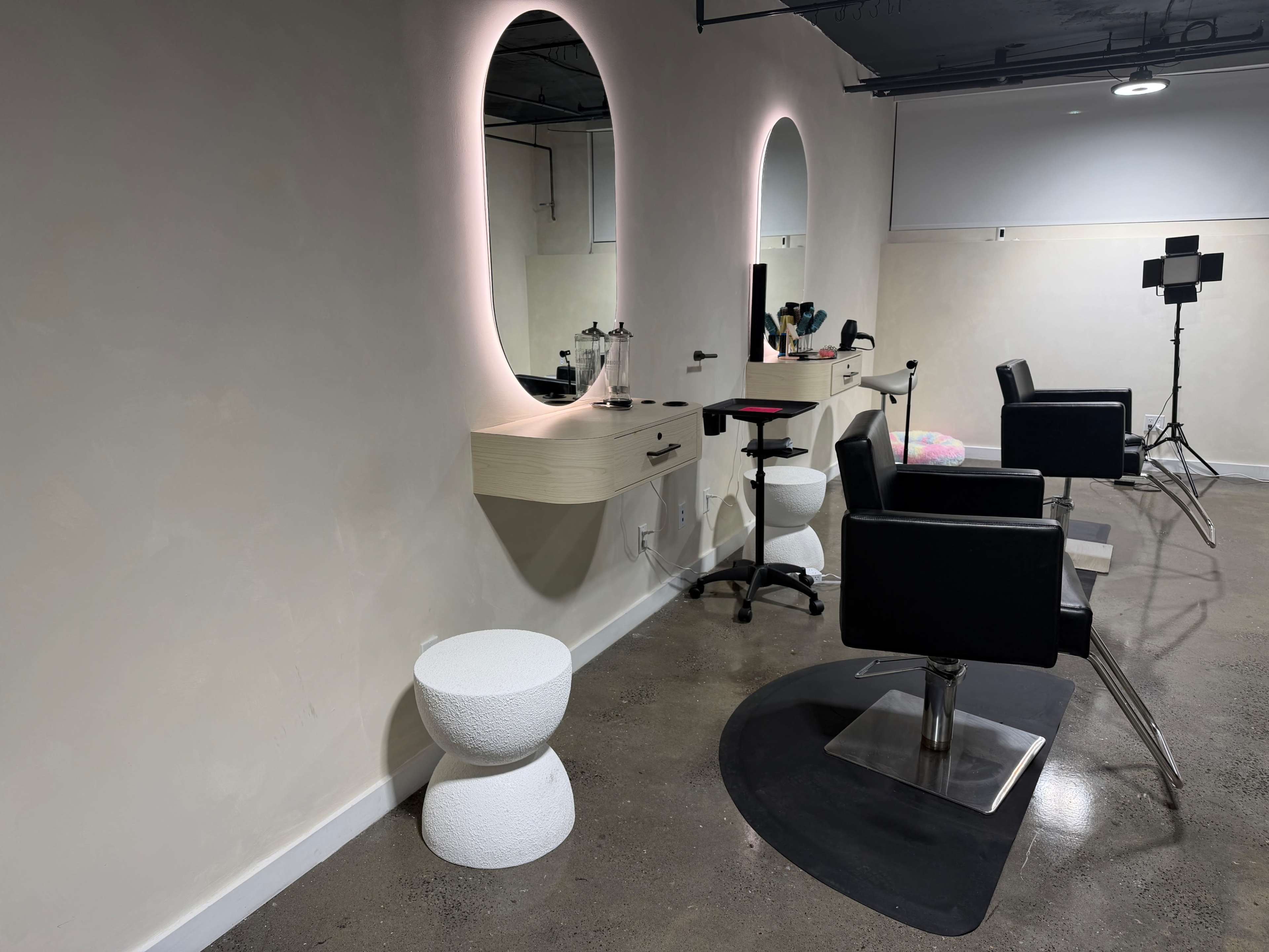 The image shows a modern hair salon interior with two black chairs, a lighted mirror, and minimalist white furnishings.