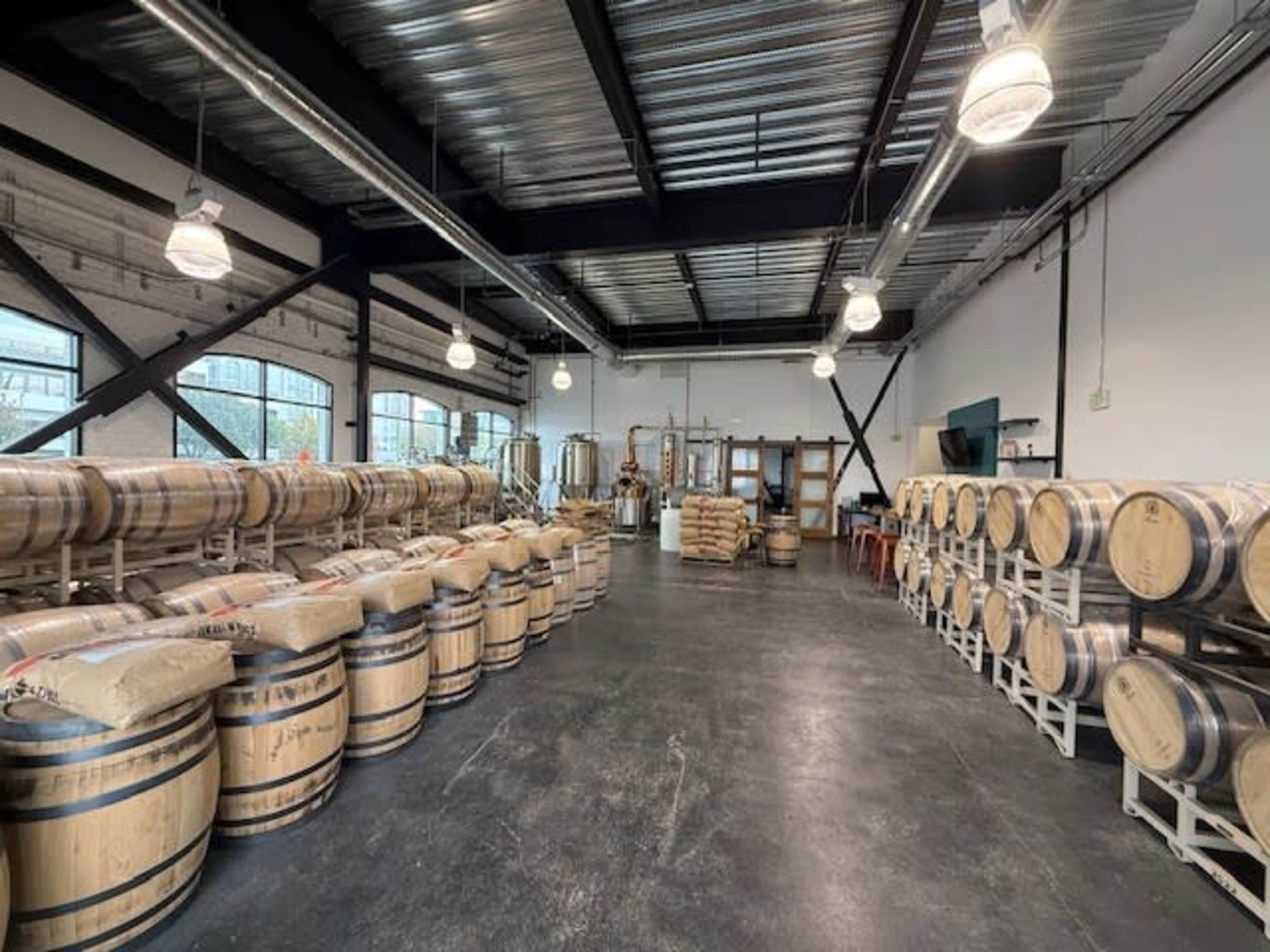 The image shows a spacious warehouse filled with wooden barrels stacked on racks, along with bags of grains and brewing equipment visible in the background.