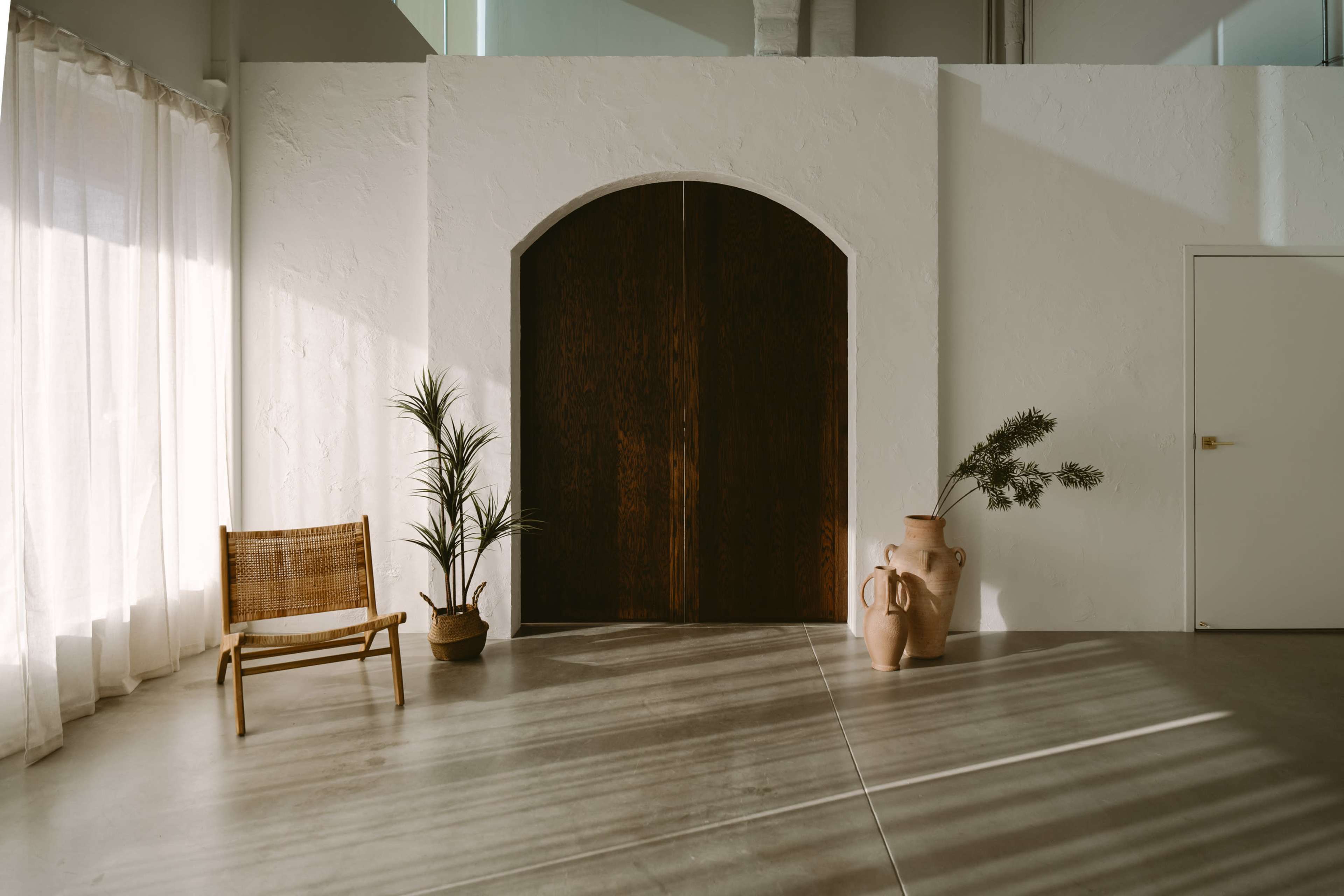 A spacious room features a large archway door flanked by a potted plant, a woven chair, and two ceramic vases, with sunlight casting shadows on the floor.