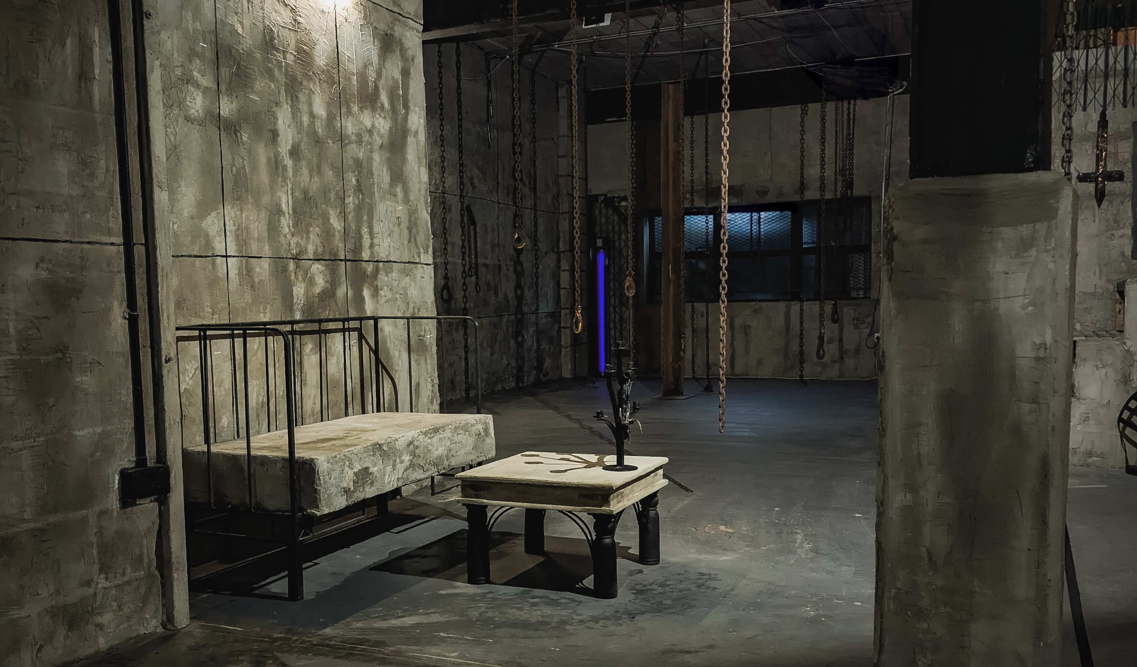 The scene features a dimly lit, industrial space with concrete walls, a bed frame, a wooden table, and hanging chains from the ceiling.