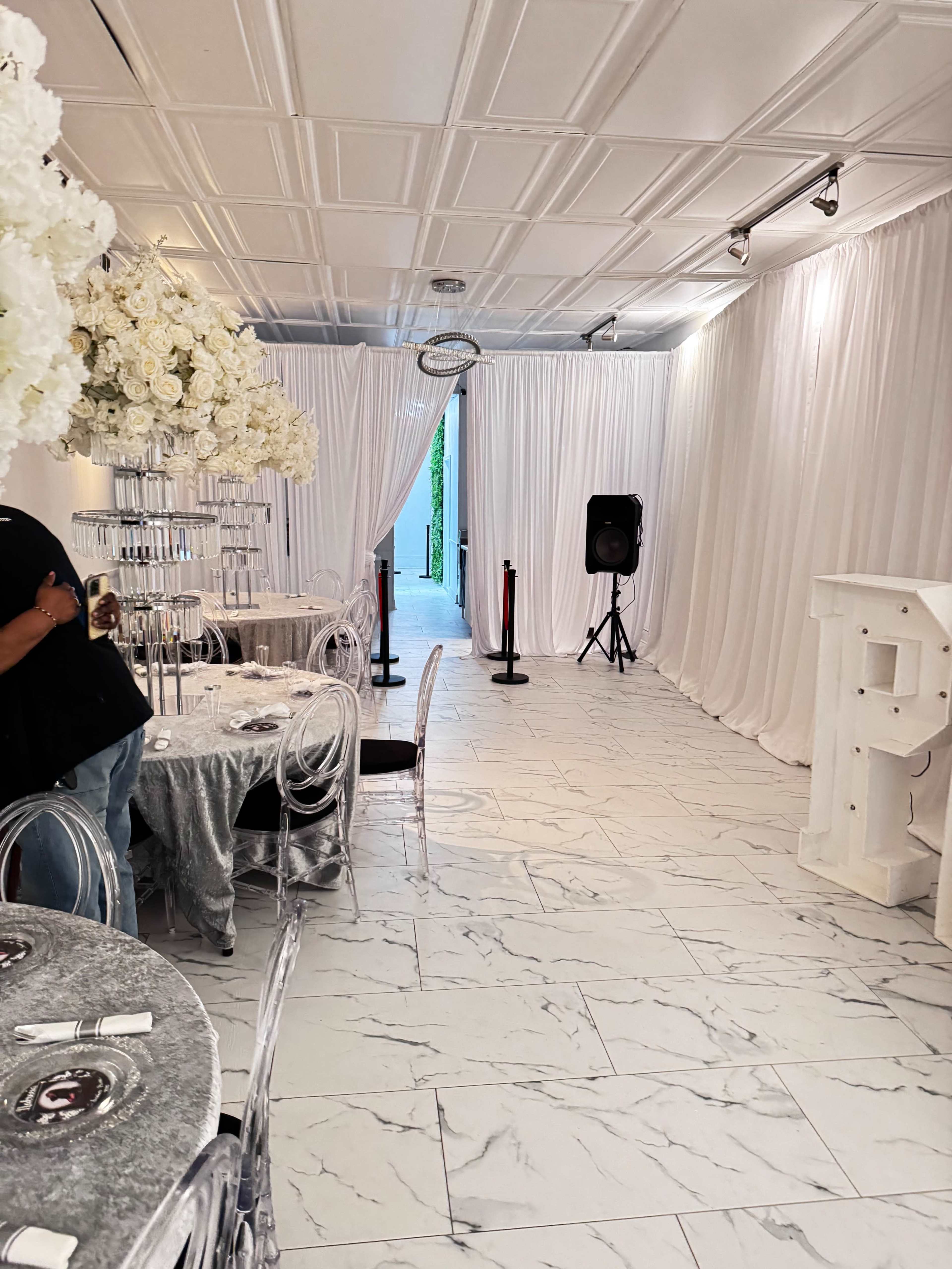 The image shows a spacious event room with white draped walls, a large floral centerpiece, clear acrylic chairs, and a speaker set up at one end.