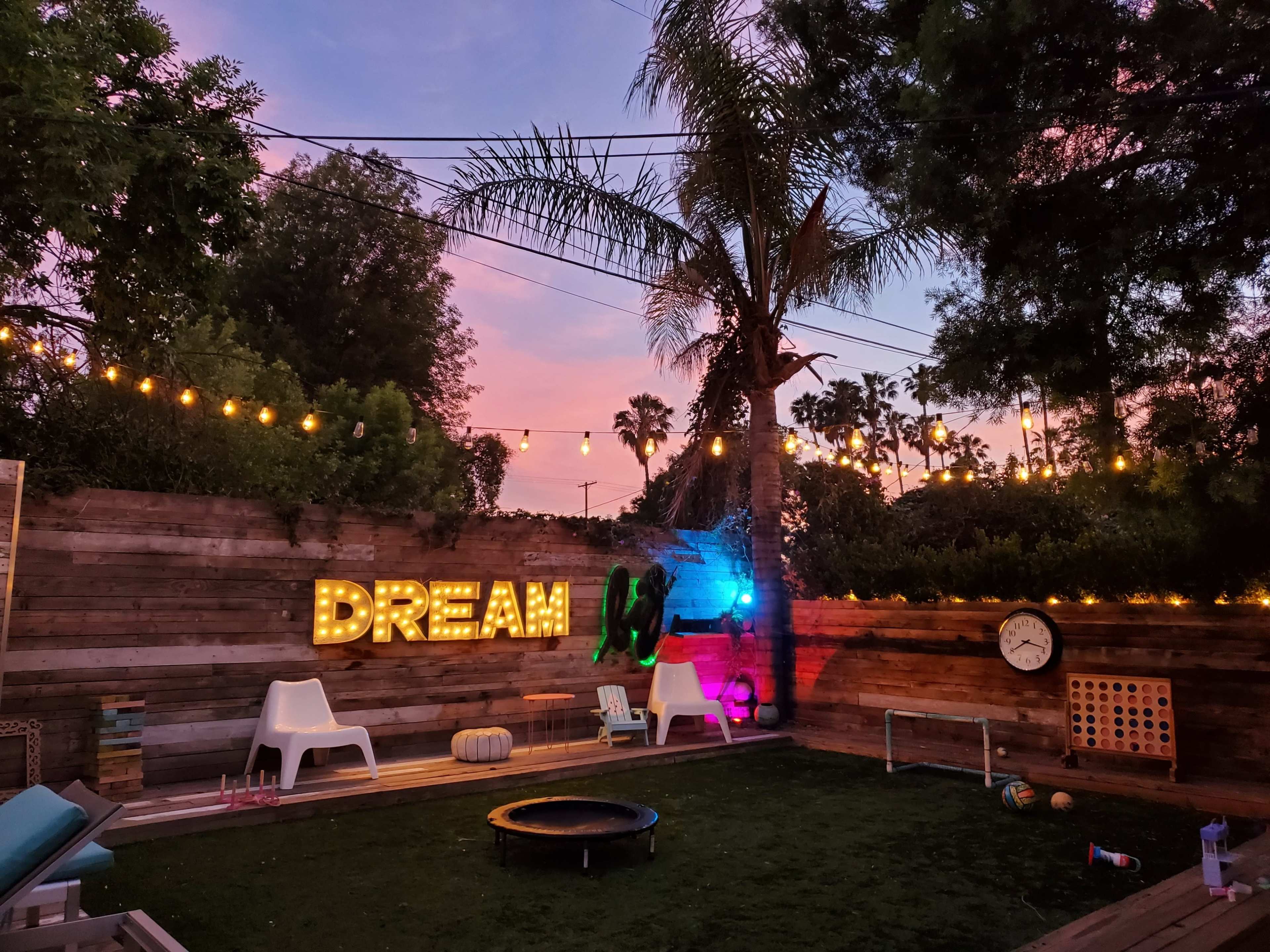 The image shows a backyard scene with a large illuminated sign that says "DREAM," surrounded by string lights, a trampoline, and various outdoor furnishings against a colorful sunset sky.