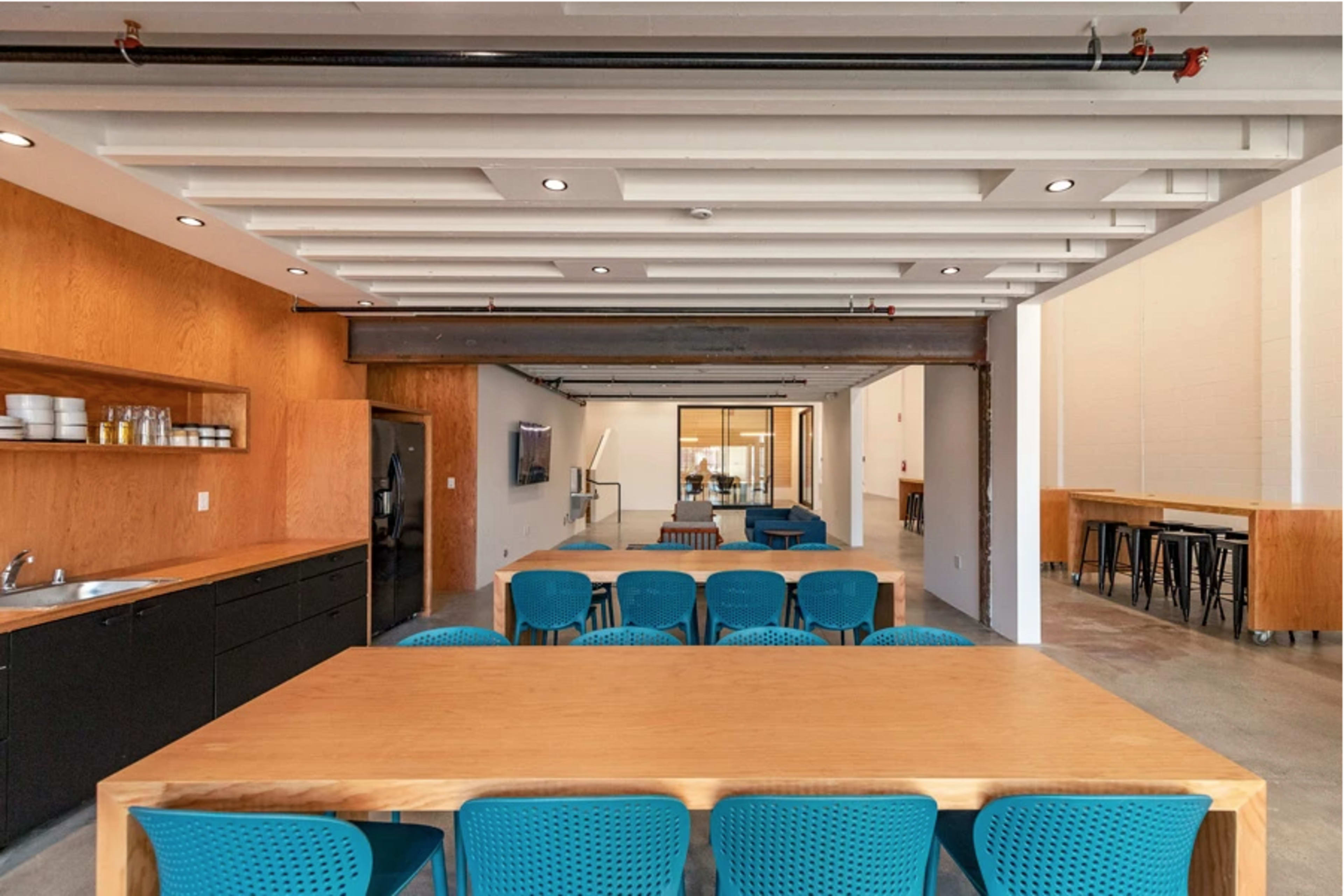 A spacious modern office area features a large wooden table with turquoise chairs, a kitchenette, and an open layout with soft lighting.