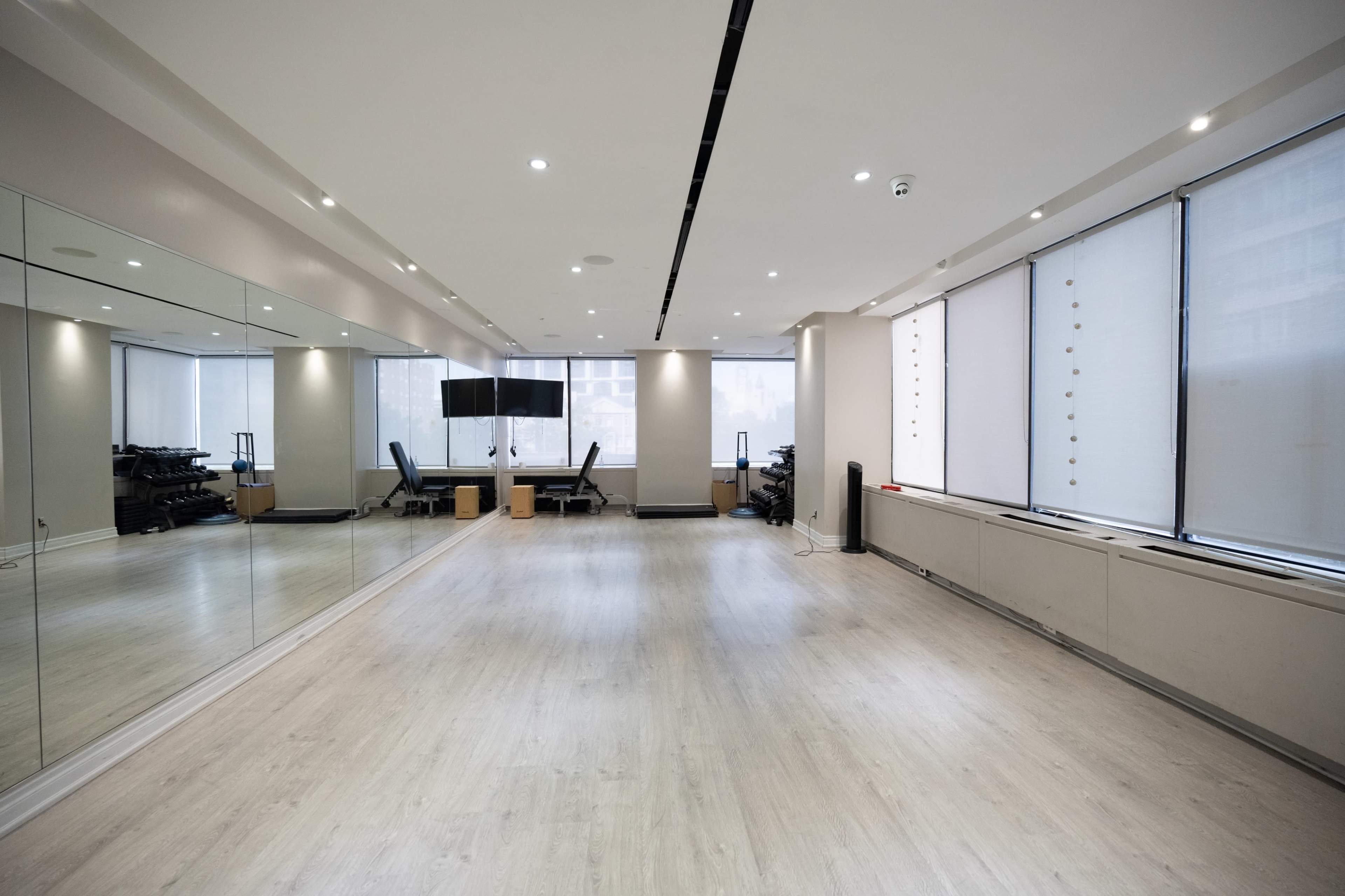 A spacious, well-lit room features a mirrored wall, wooden flooring, and modern office furniture, including chairs and desks.