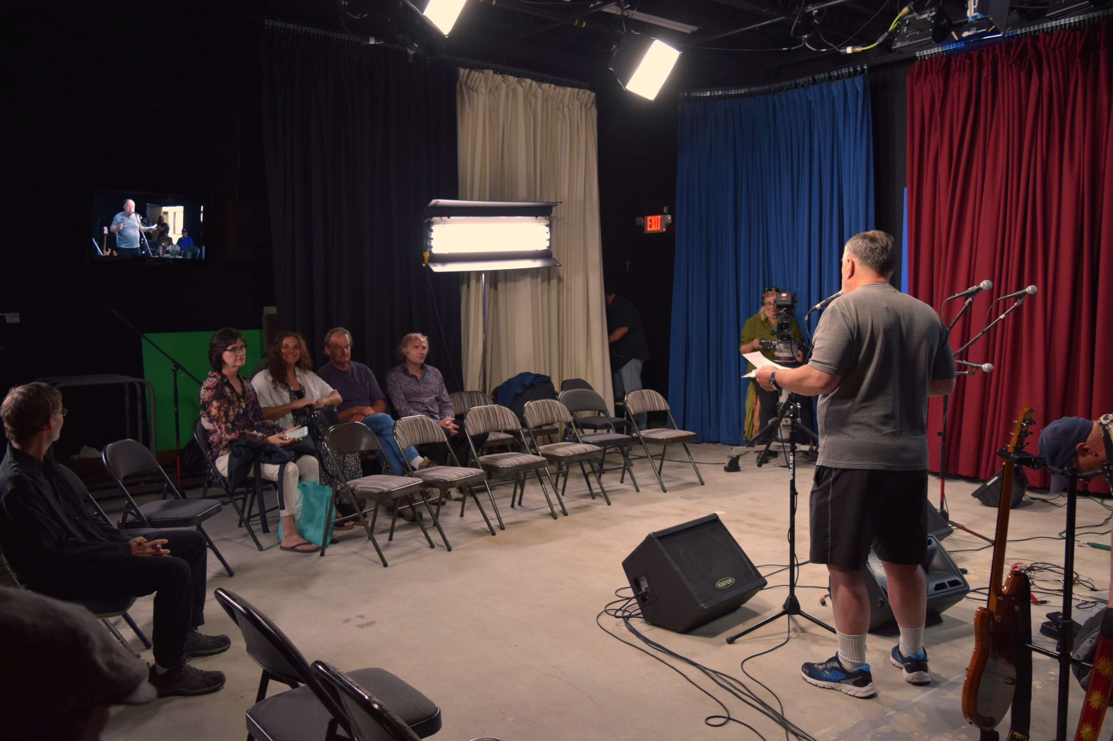 A man stands on stage speaking to an audience seated on folding chairs in a studio with red, blue, and white curtains.