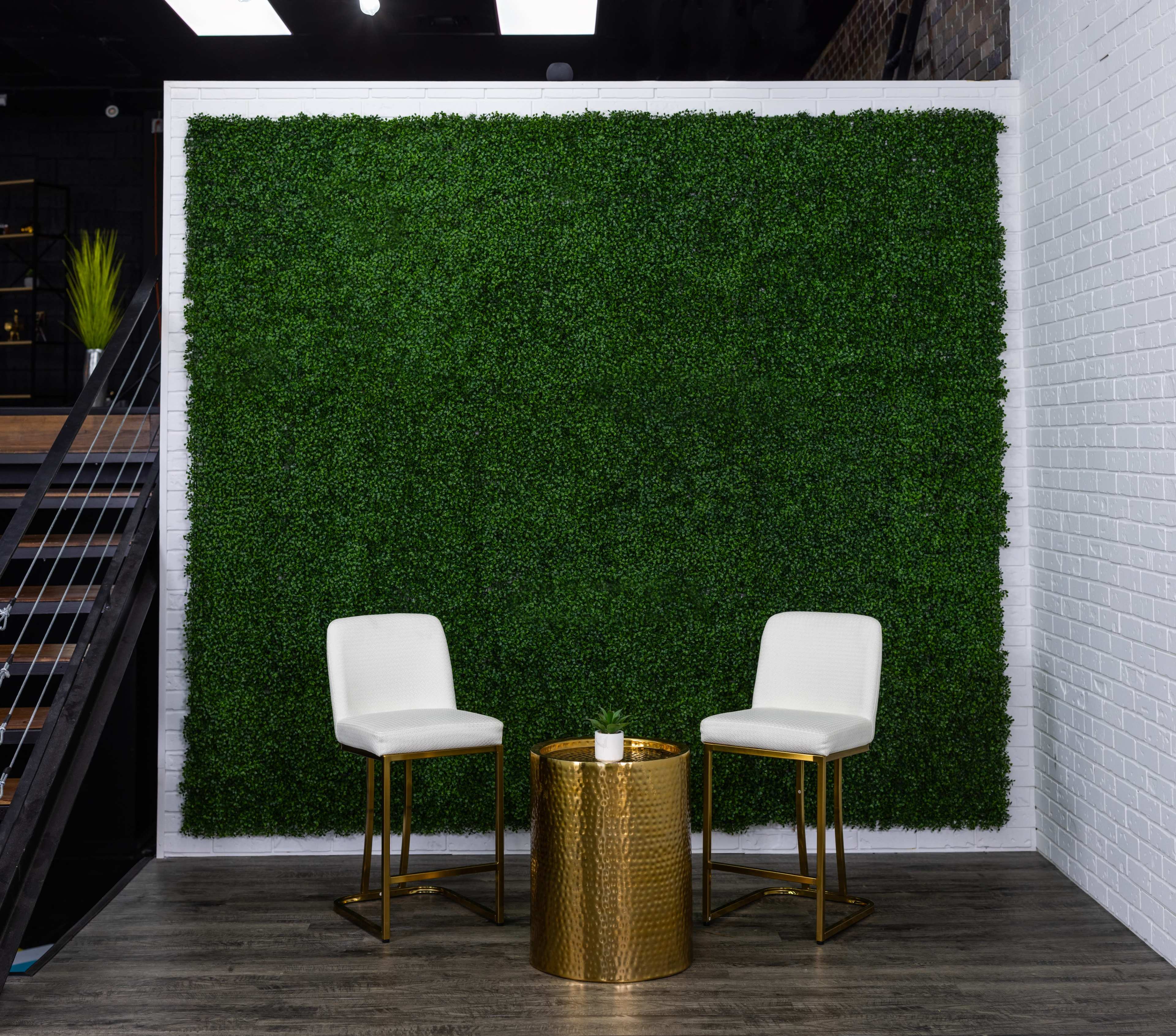 A green artificial grass wall with two white chairs facing a gold circular table.