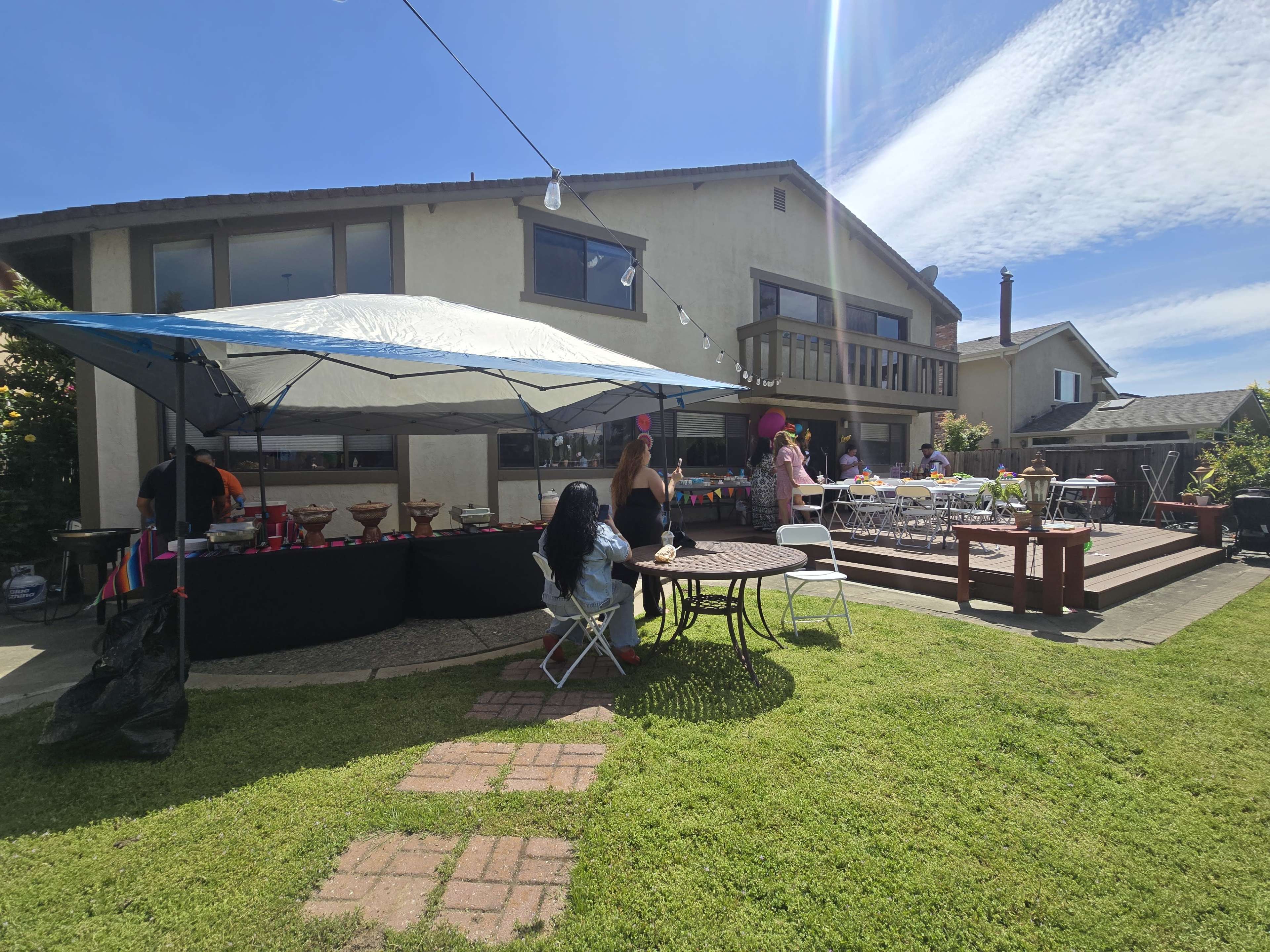 A backyard party is set up with tables, chairs, and a food service area under a tent, alongside a two-story house.
