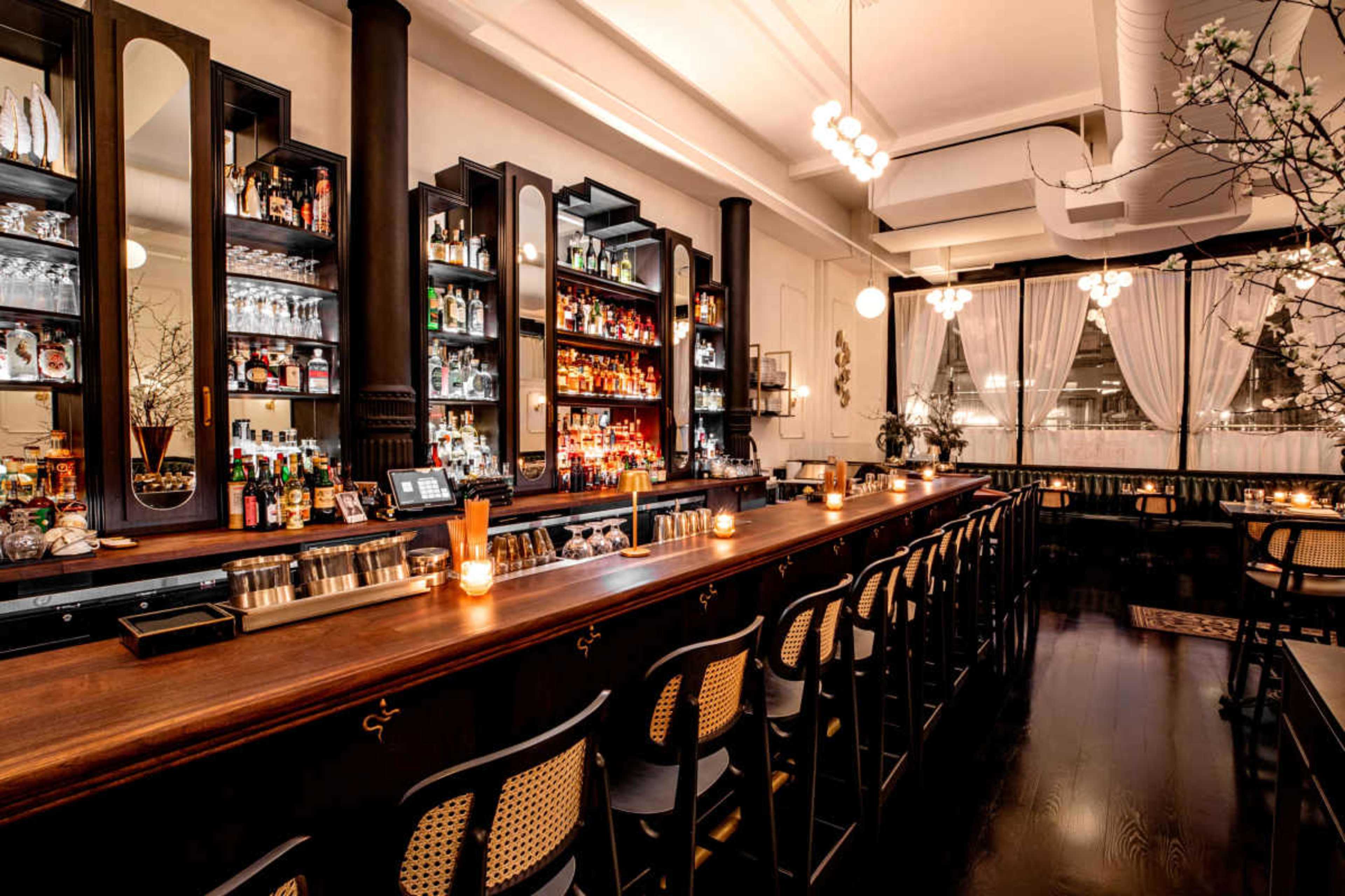 A well-lit bar interior features a wooden counter, shelves stocked with various bottles, and stylish seating along the length of the bar.