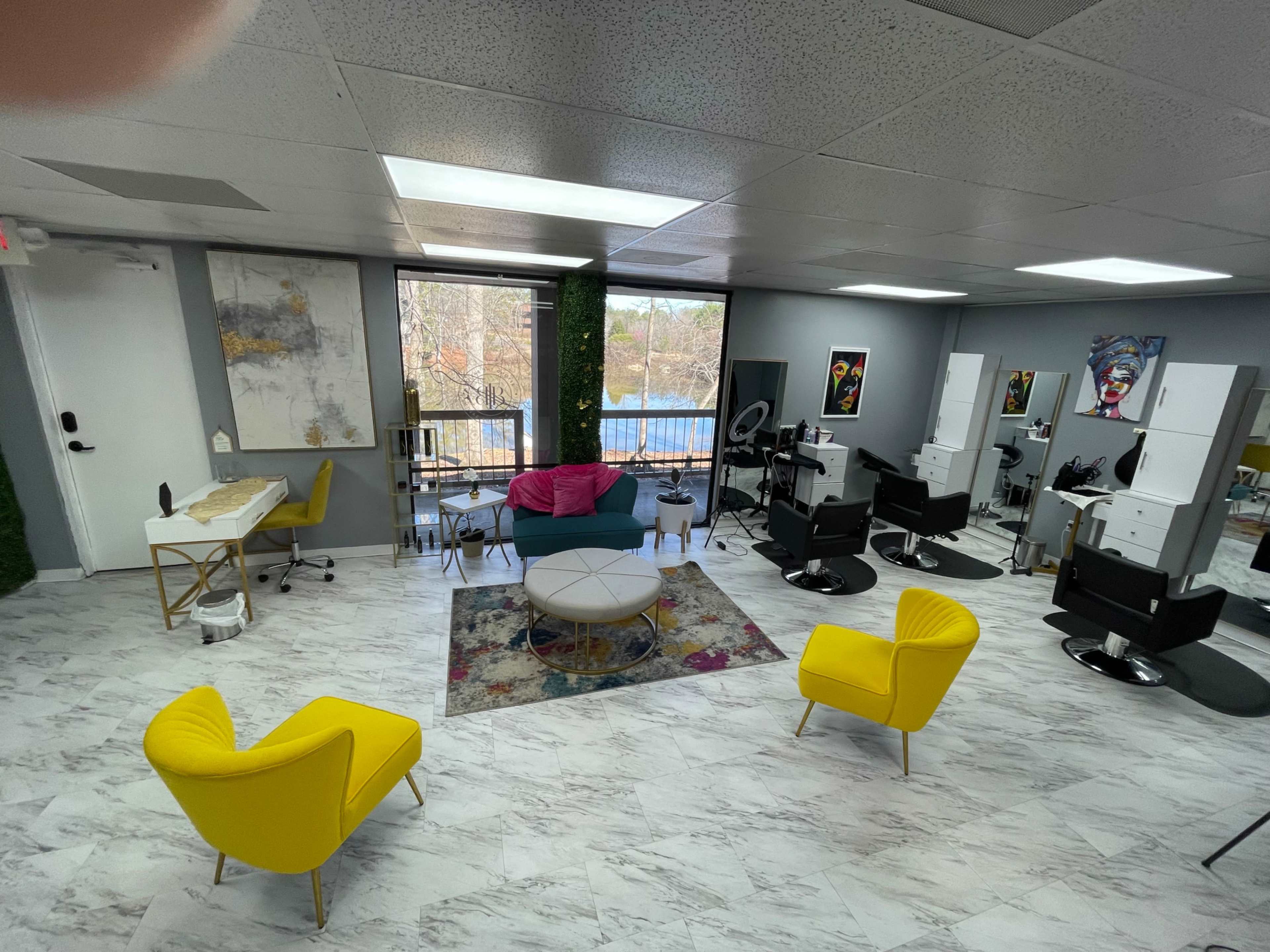 A modern hairstyling salon with yellow chairs, a styling station, a seating area, and large windows providing a view of the outdoors.