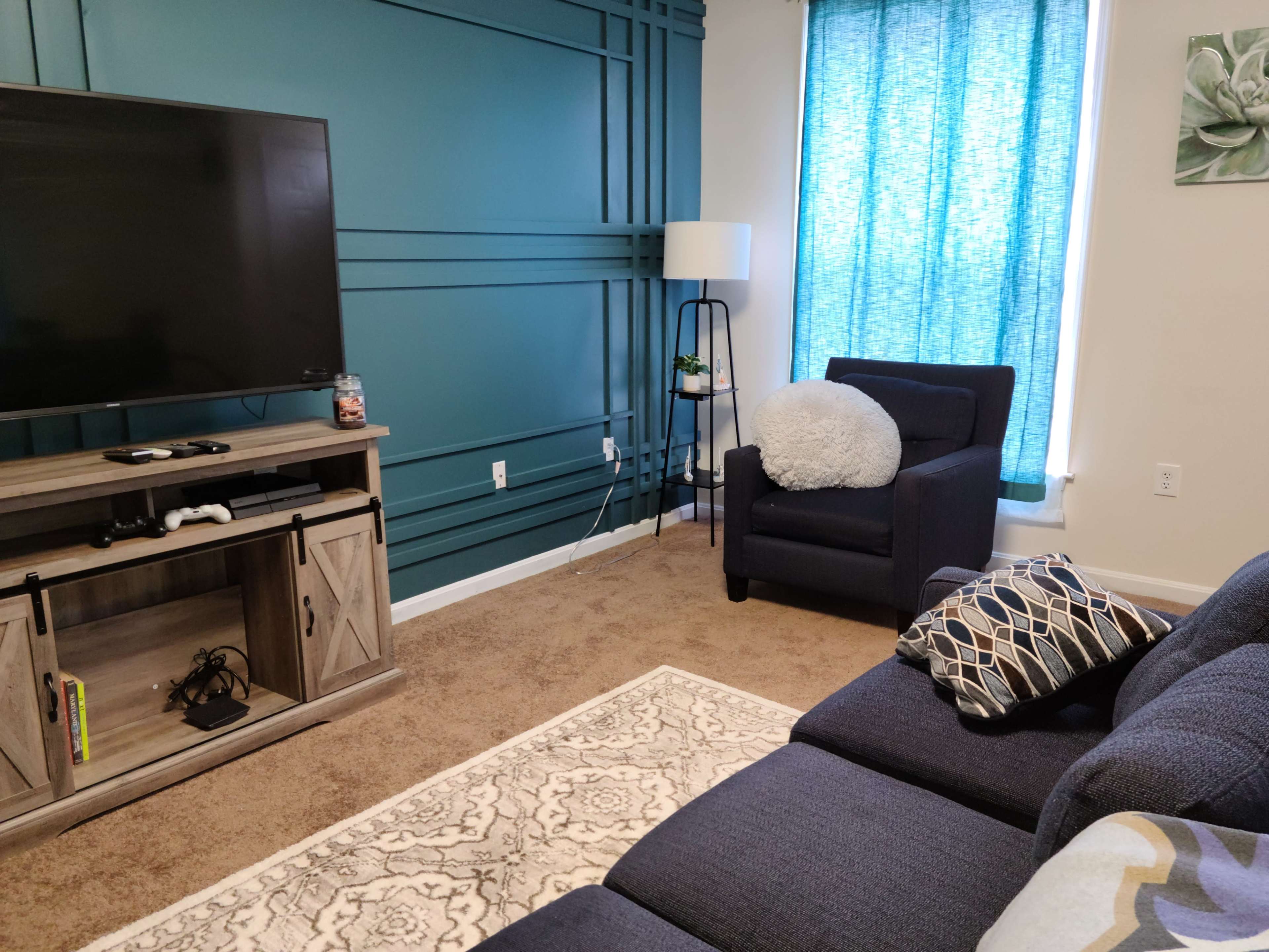 A living room with a dark blue accent wall, a large television mounted on a wooden media console, and a seating area featuring a dark blue couch and a single armchair near a window with light blue curtains.