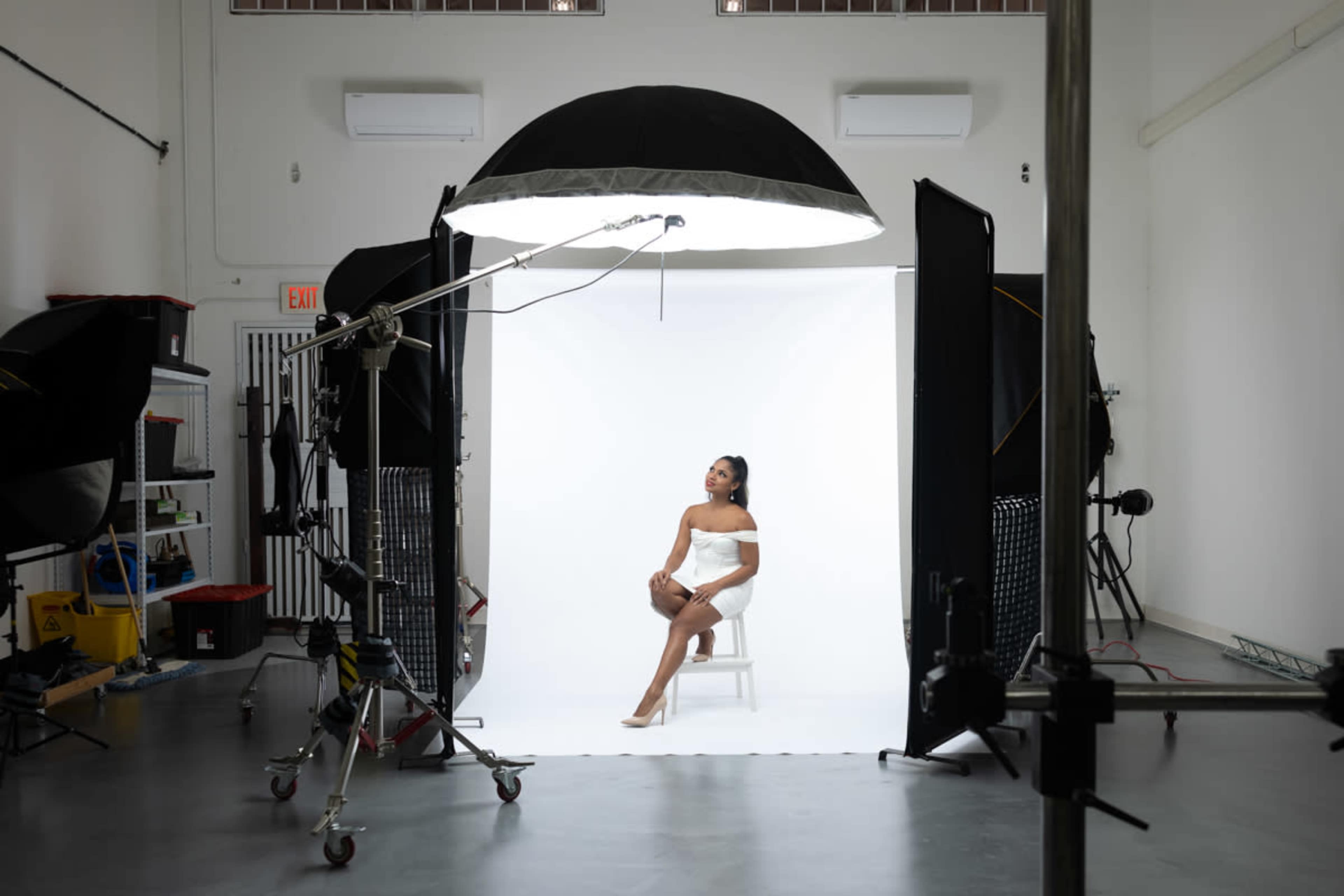 A model poses on a chair against a white backdrop in a photography studio equipped with various lighting and support equipment.