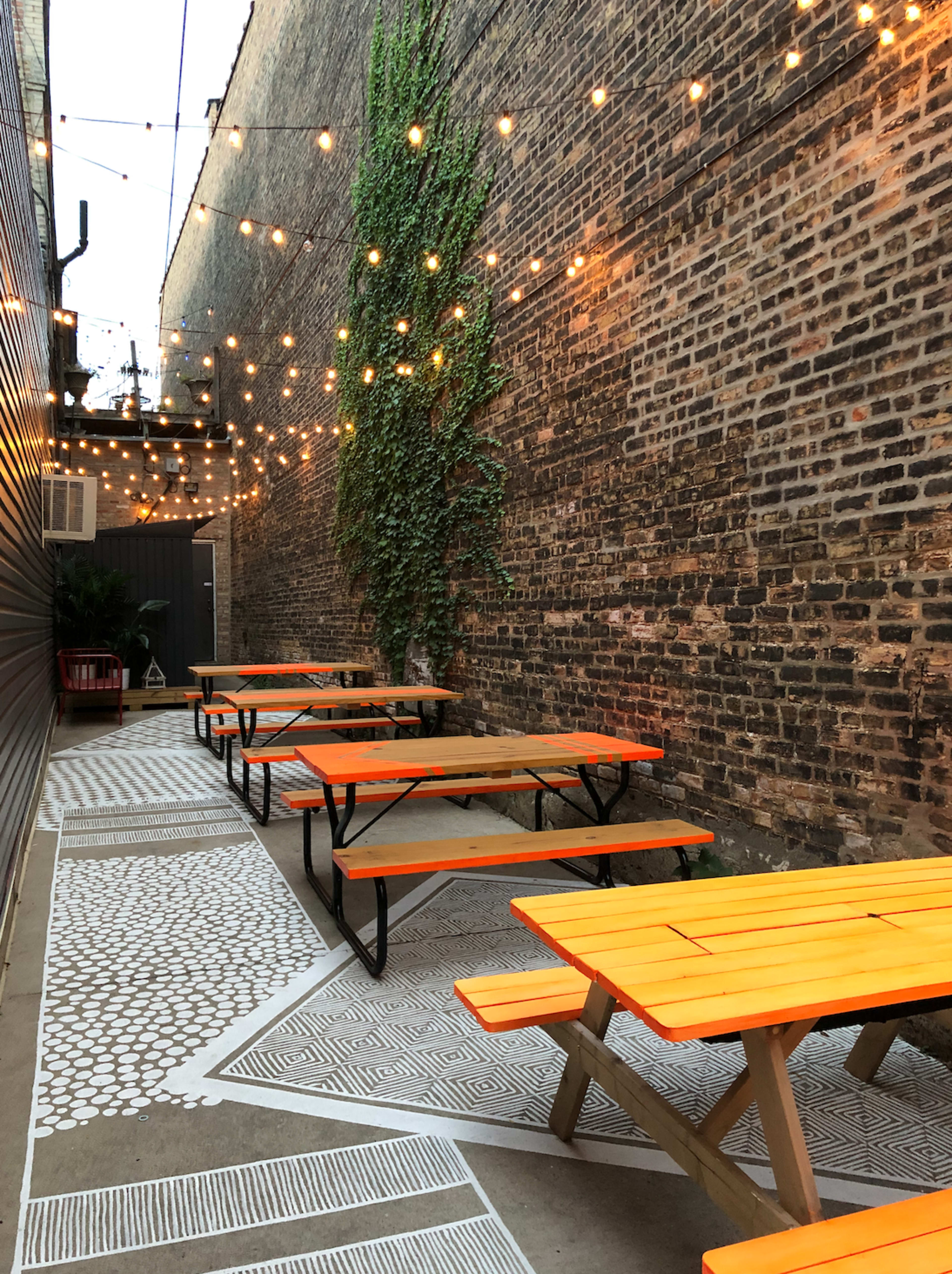 A narrow outdoor seating area lined with wooden picnic tables painted orange under string lights and a brick wall.