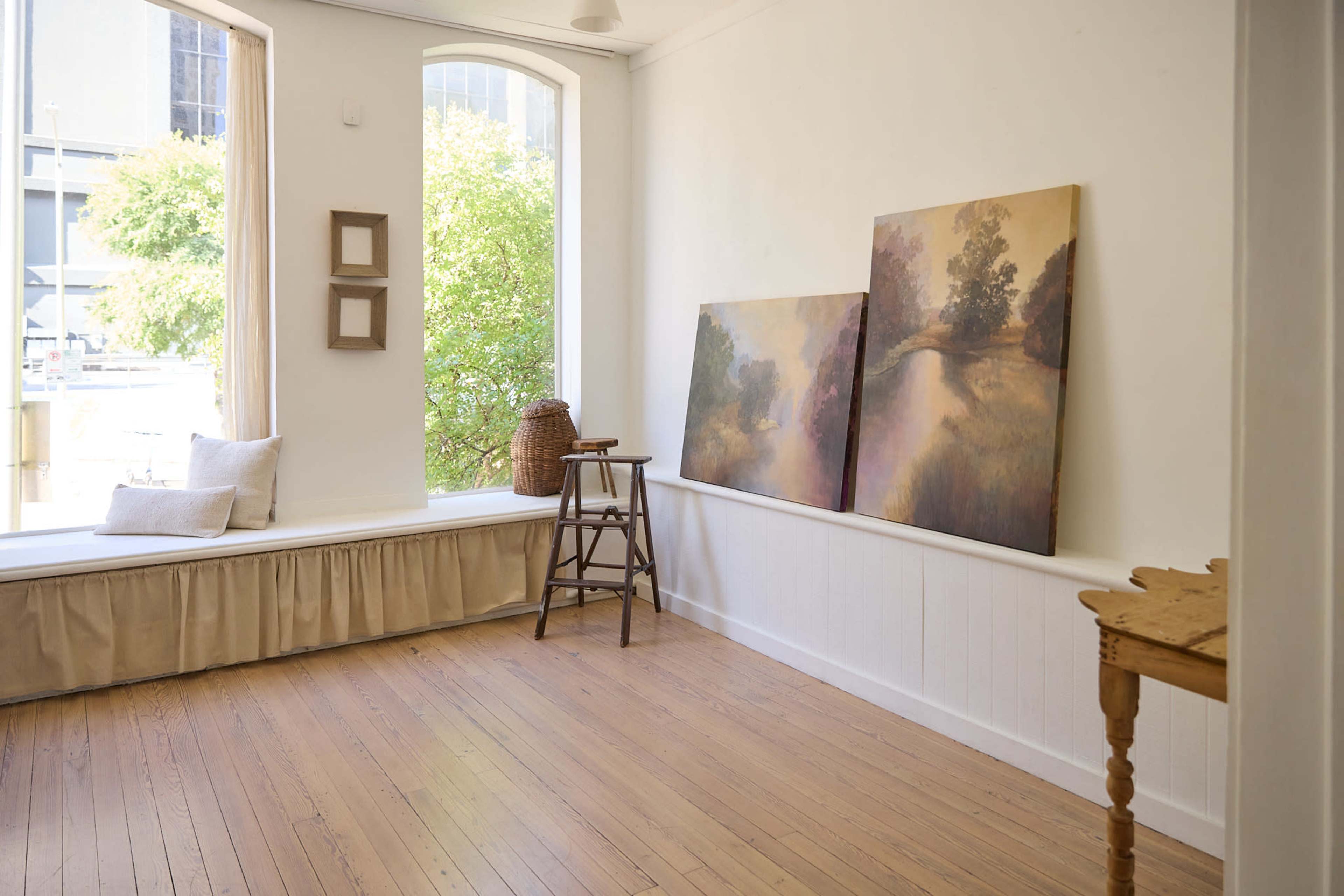 The image shows a bright, minimalist room with wooden flooring, featuring two landscape paintings on the wall and a small wooden table near a window.