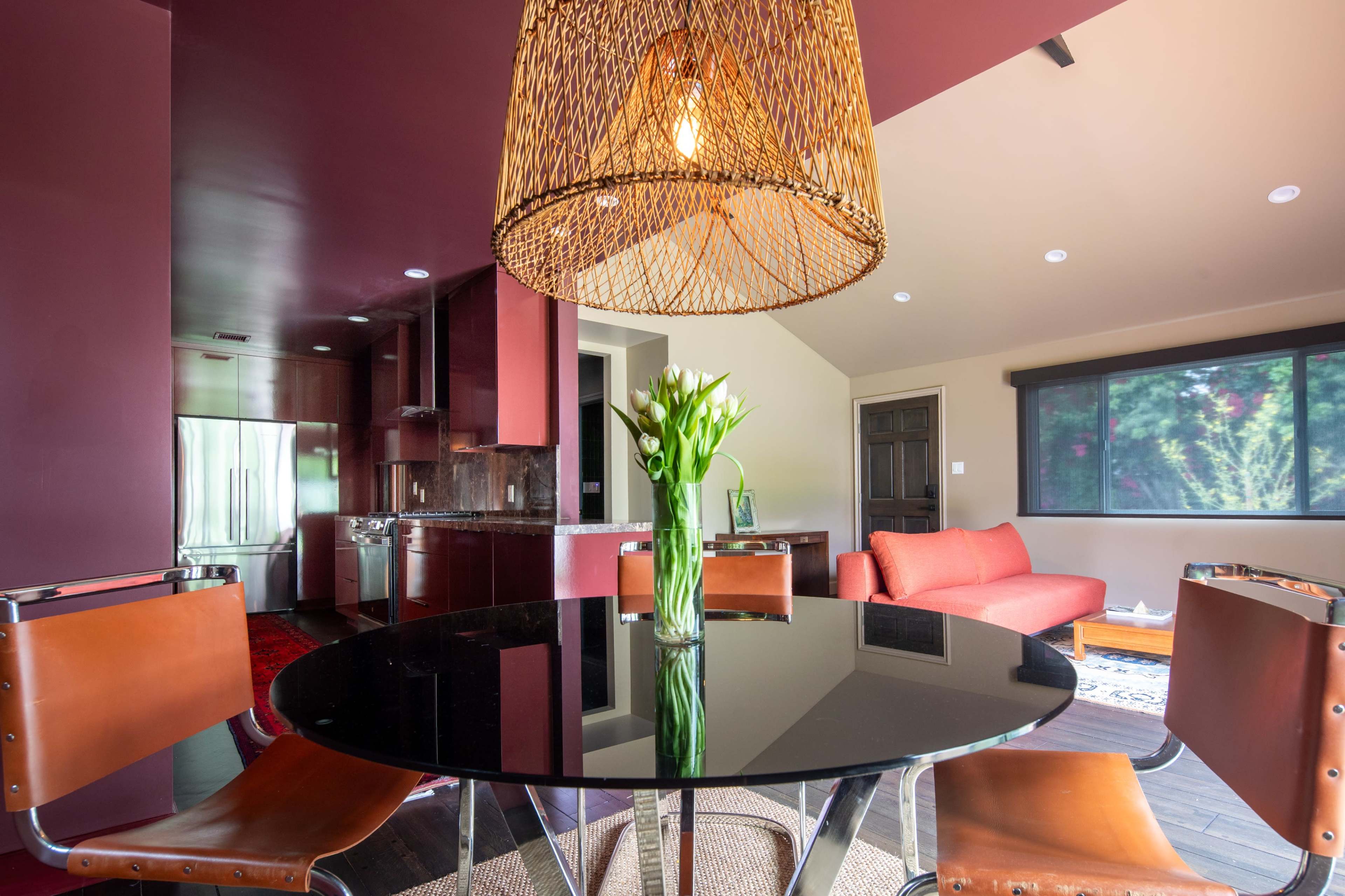 A modern dining area features a round black table with four leather chairs, a large woven pendant light, and a vase of tulips in the center.