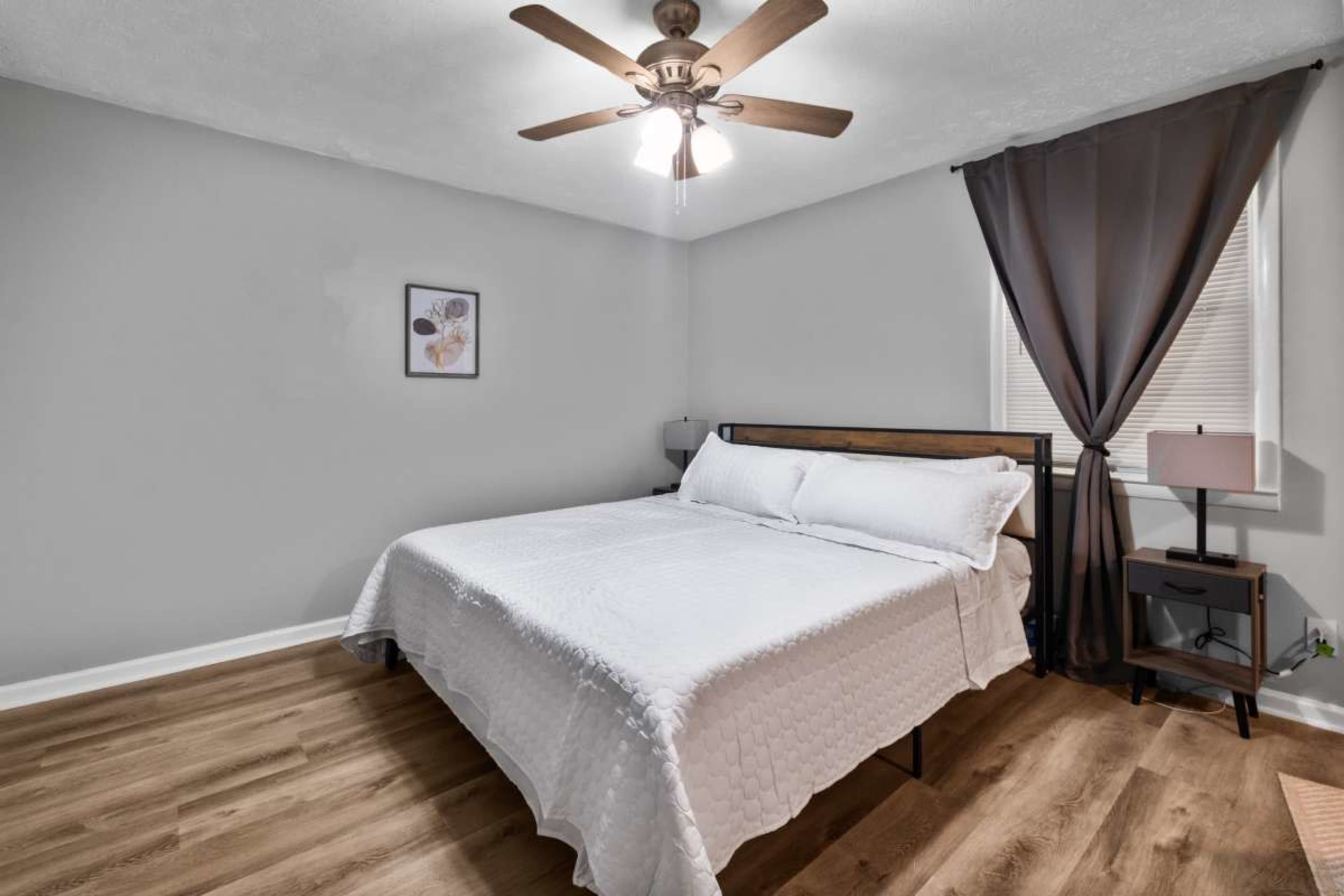 The image shows a simple bedroom featuring a queen-sized bed with a white quilt, a ceiling fan, and a nightstand with a lamp.