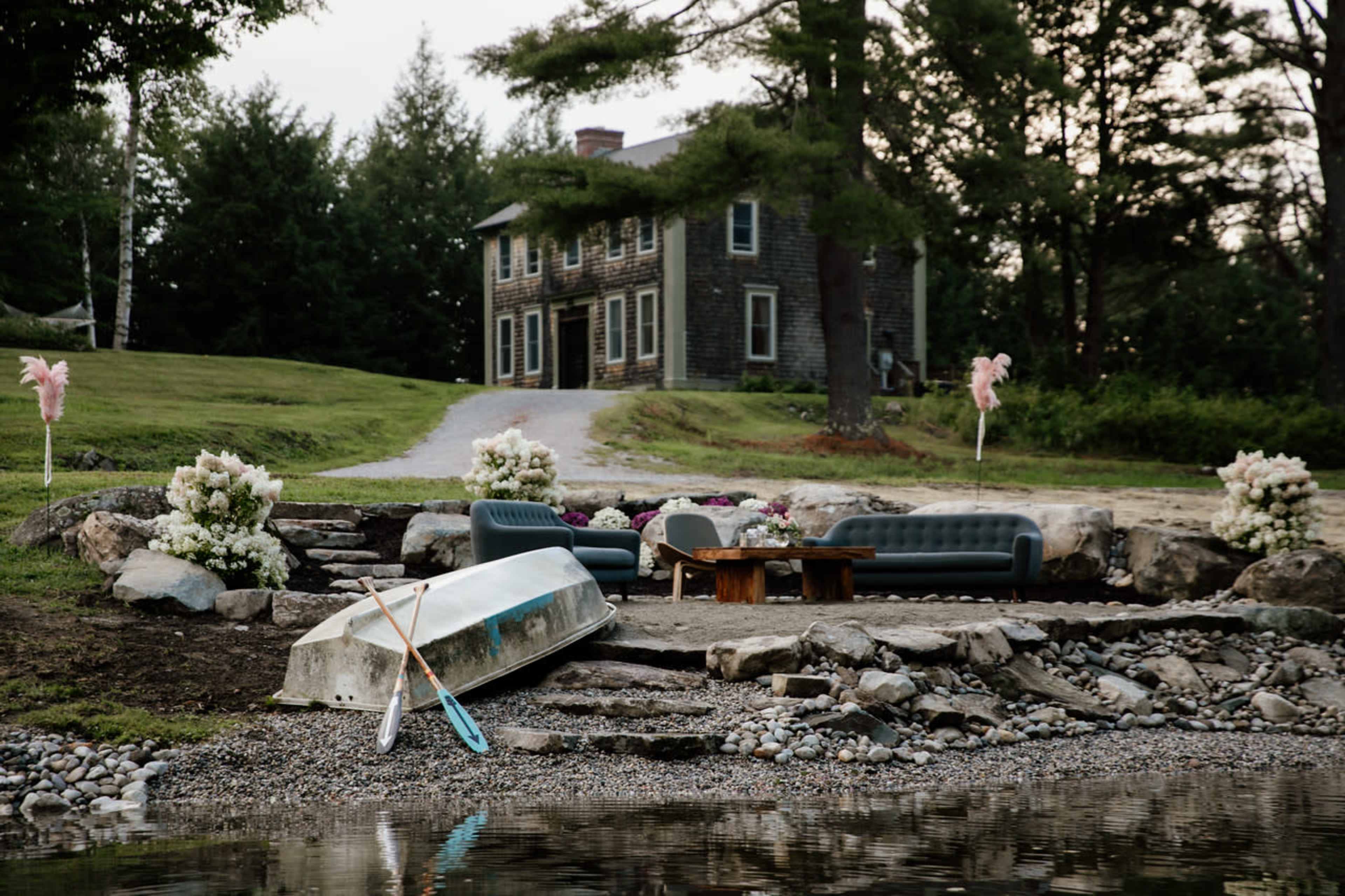A rustic scene features a lakeside gathering area with two sofas, a coffee table, a rowboat, and flower arrangements, set against a stone house and tree line.