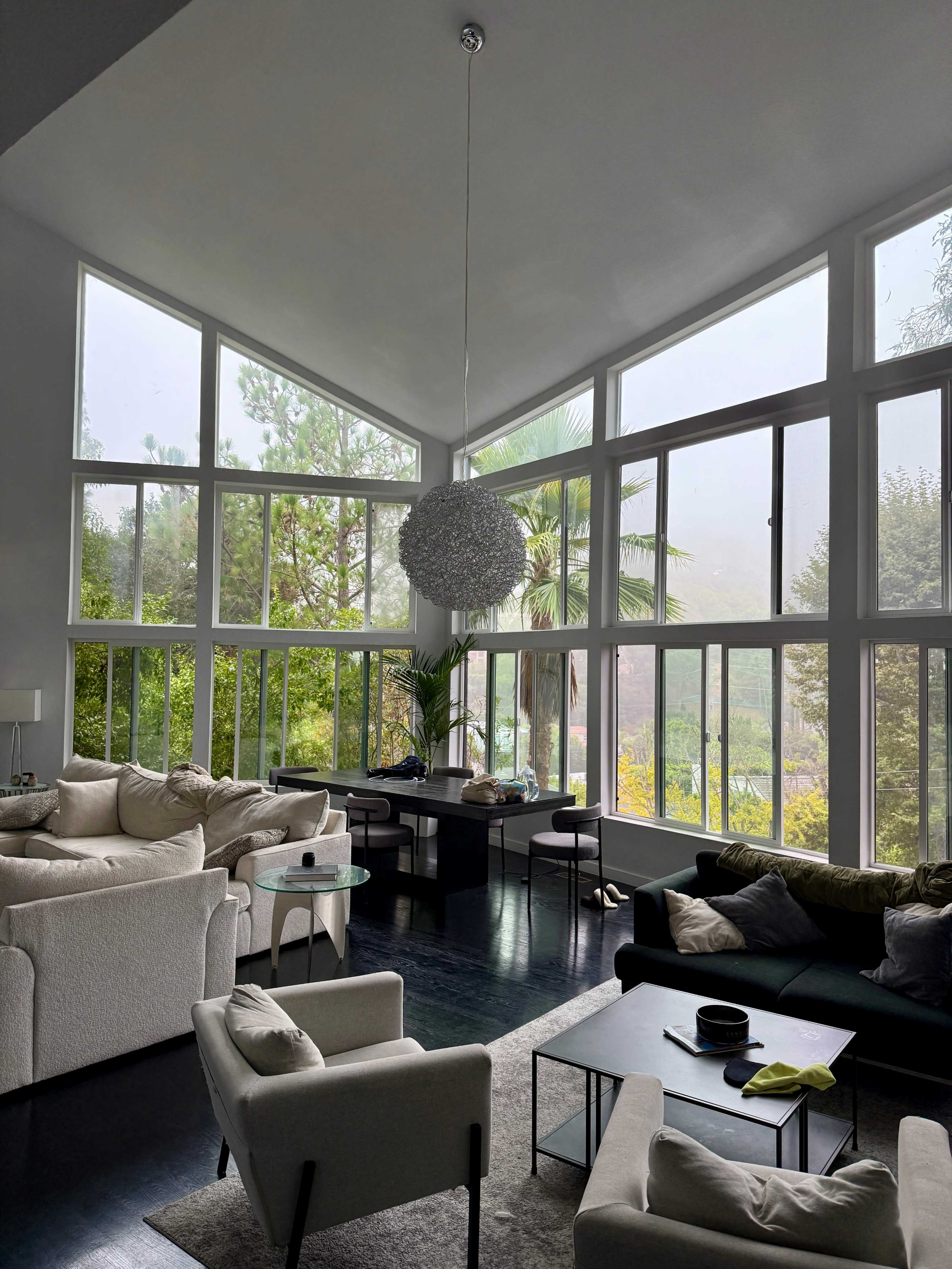 A spacious living room features large windows, a modern pendant light, and a mix of white and dark furniture, with views of greenery outside.