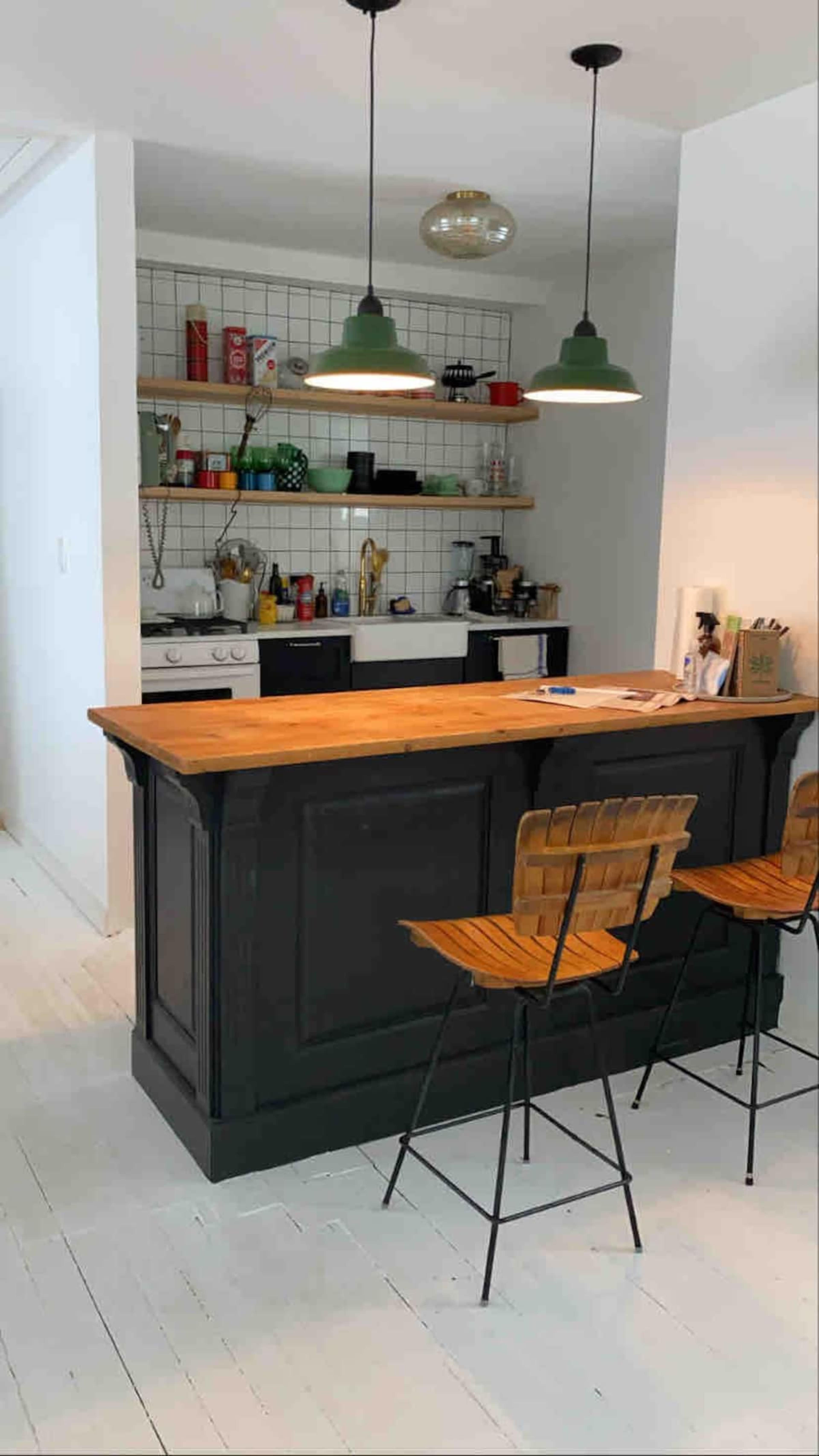 A modern kitchen features a dark wood bar with two wooden stools, open shelves displaying kitchen items, and pendant lights hanging above.
