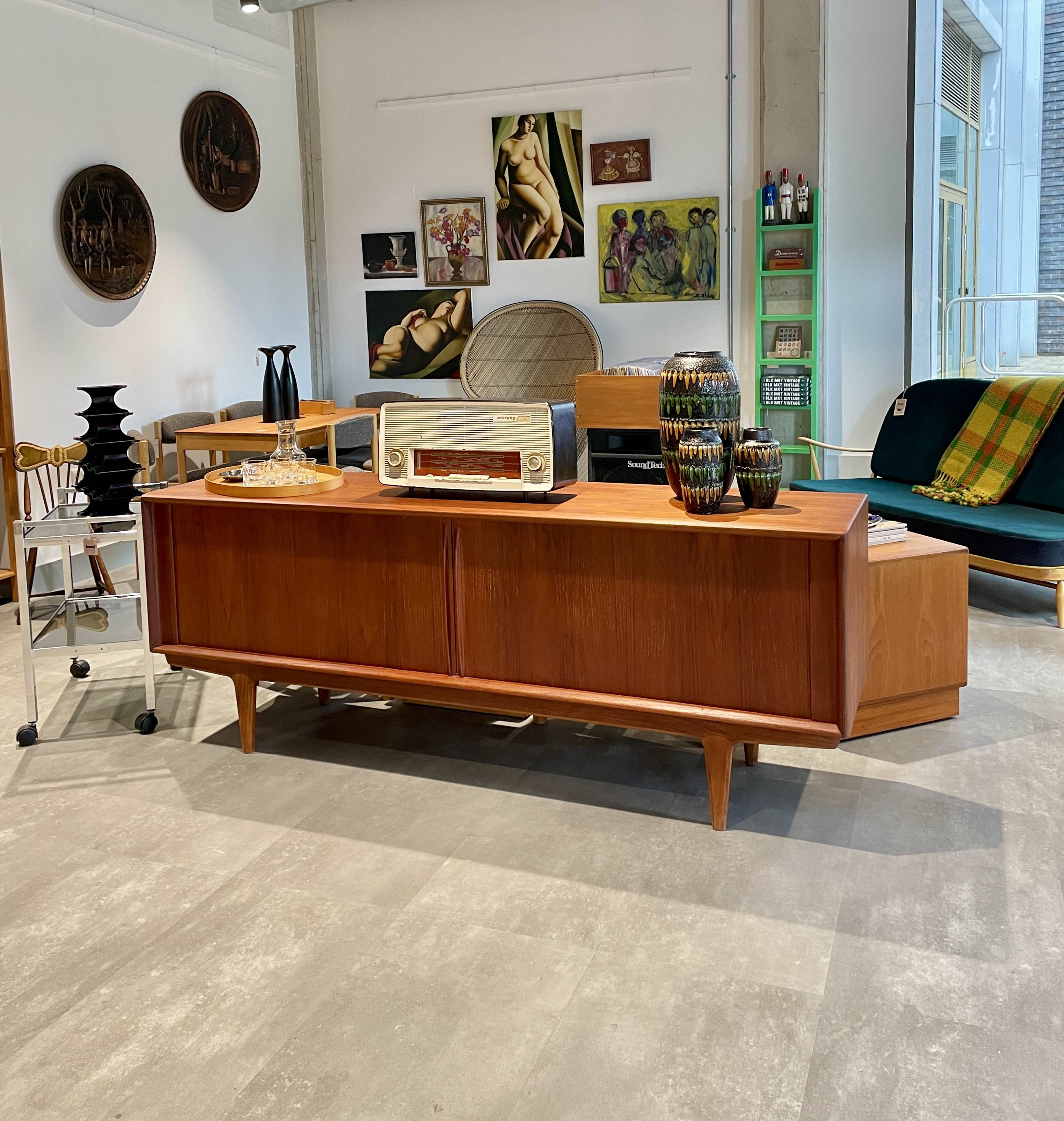 The image shows a modern interior with a wooden sideboard featuring a vintage radio, decorative vases, and various artworks on the walls, alongside mid-century furniture pieces.