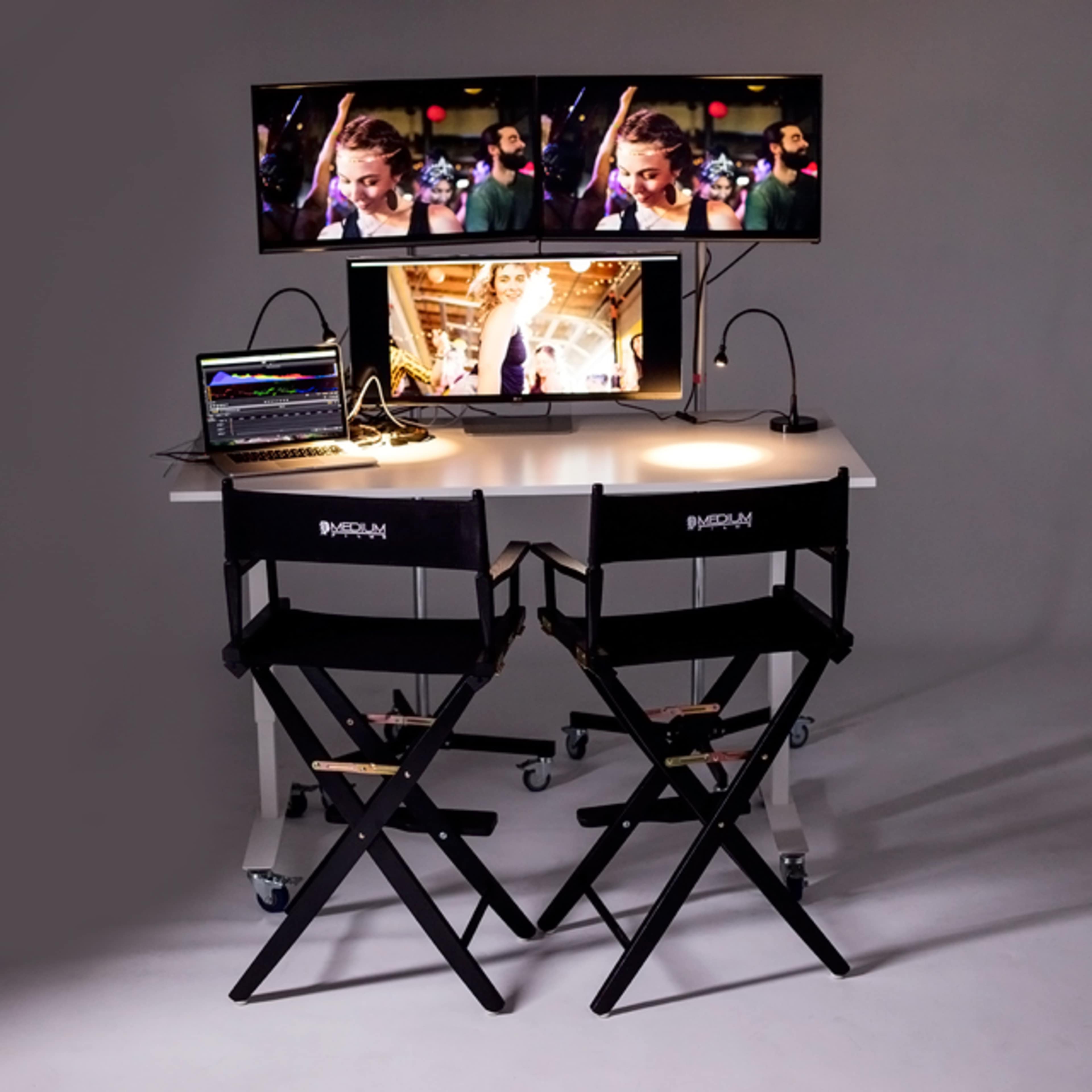 A workspace is shown featuring two monitors, a laptop, and two director's chairs on a white table, all set against a gray backdrop.