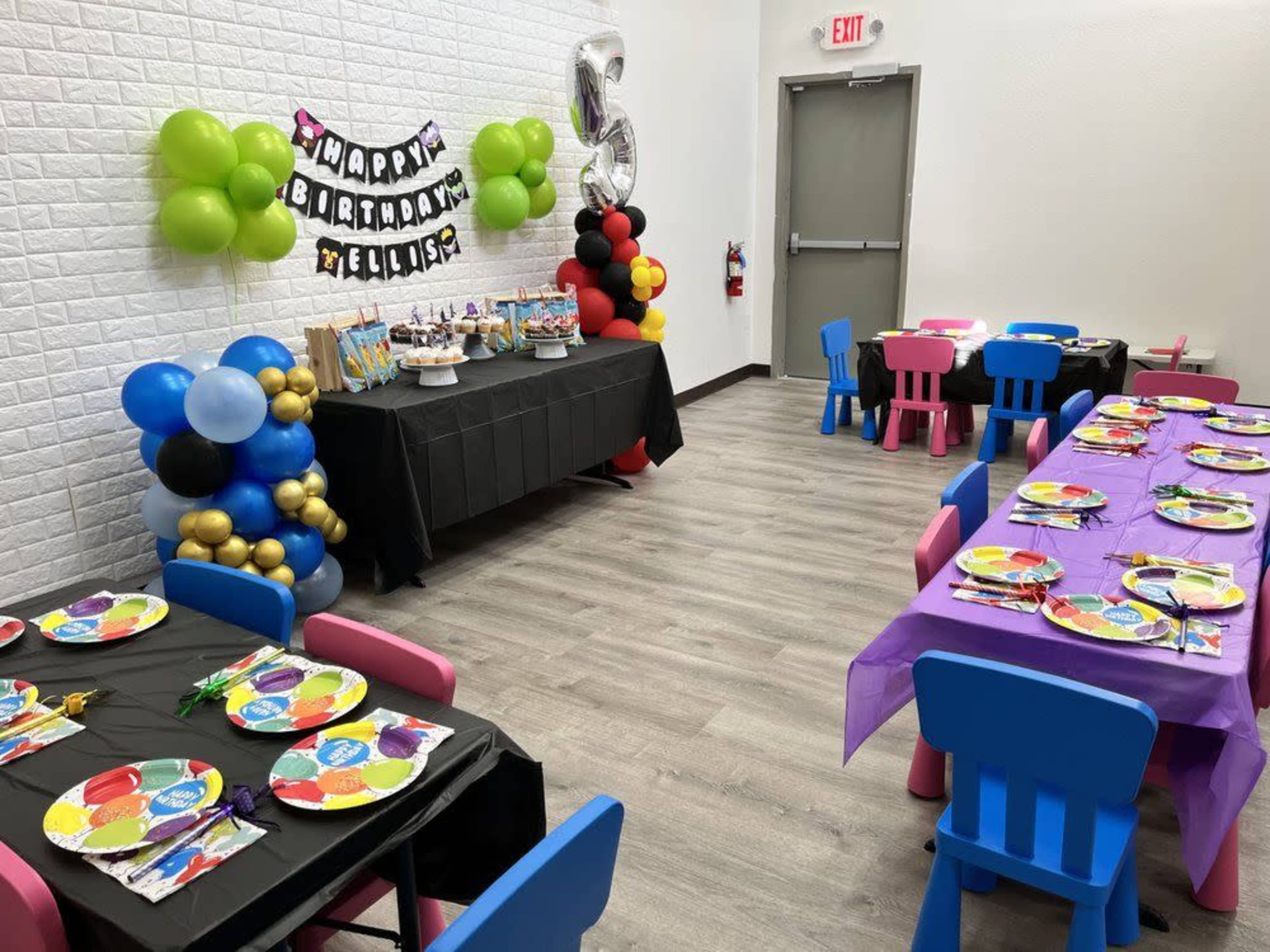 A birthday party setup features two tables adorned with colorful decorations, plates, and balloons, alongside a dessert table with treats and a banner.