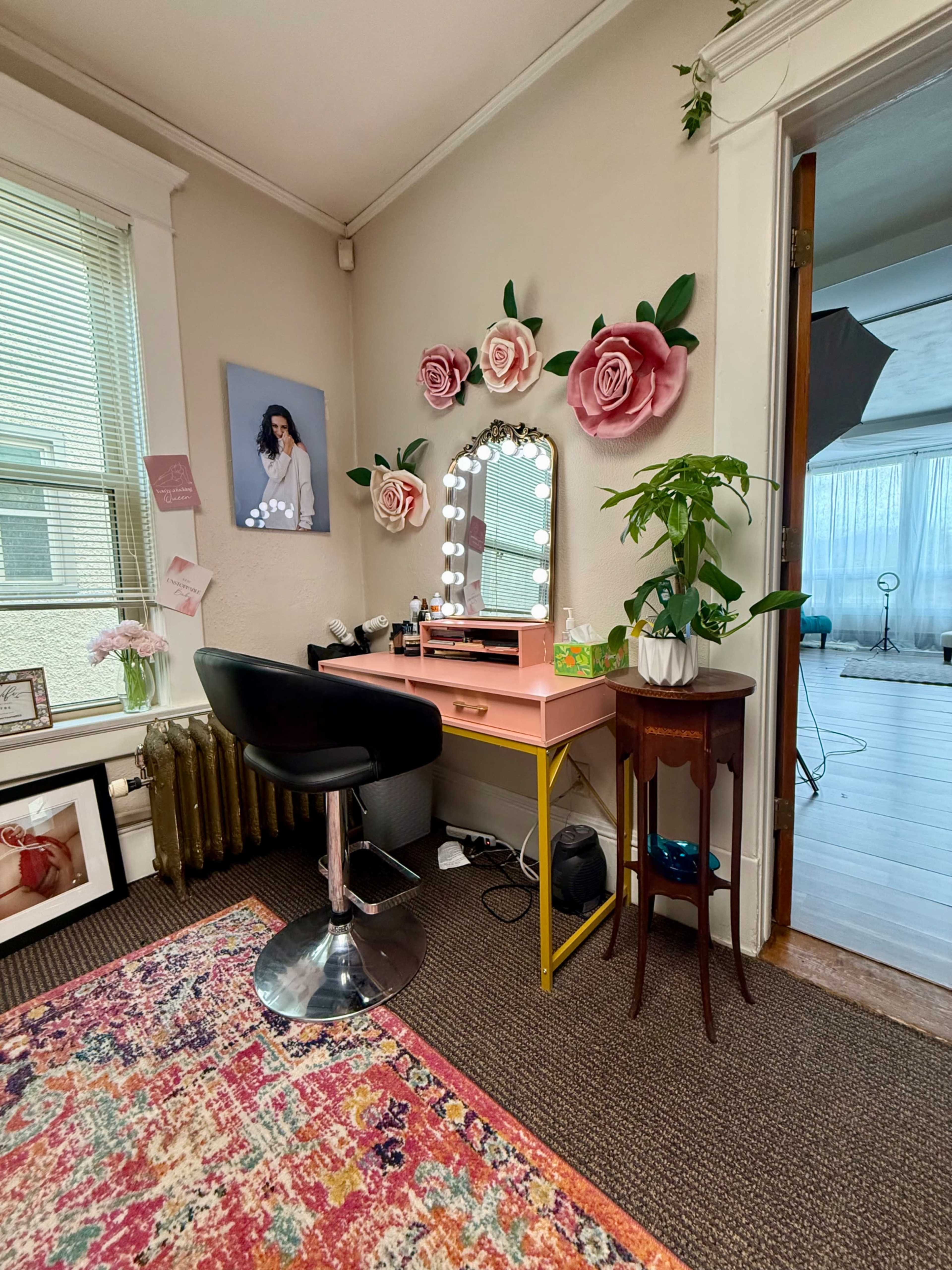 The image shows a brightly decorated vanity area with a large mirror adorned with lights, a black swivel chair, a small side table, and floral wall decor in a well-lit room.
