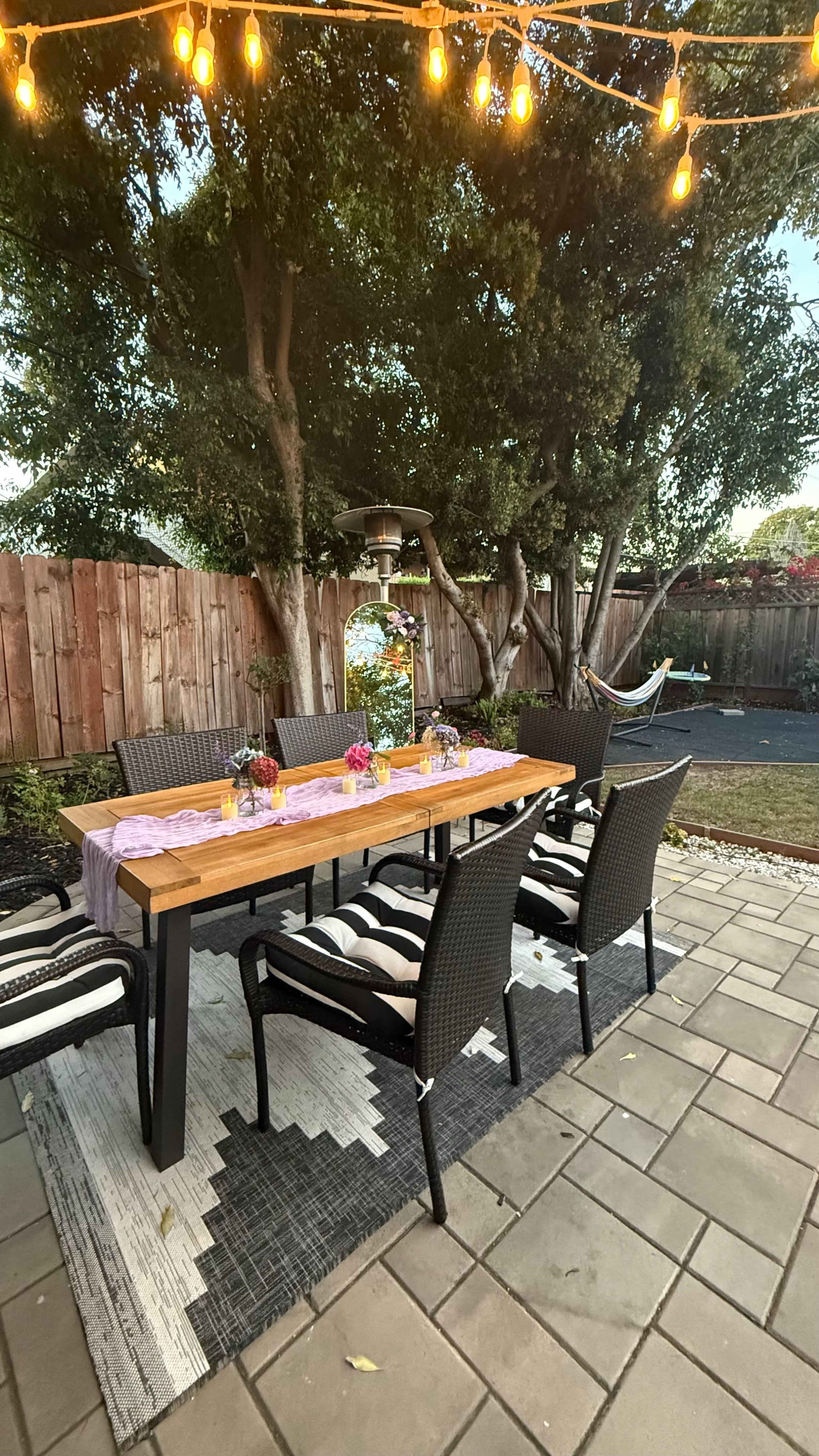 A wooden dining table surrounded by black wicker chairs is set with flower centerpieces and a pink table runner in a backyard patio with string lights and a hammock in the background.