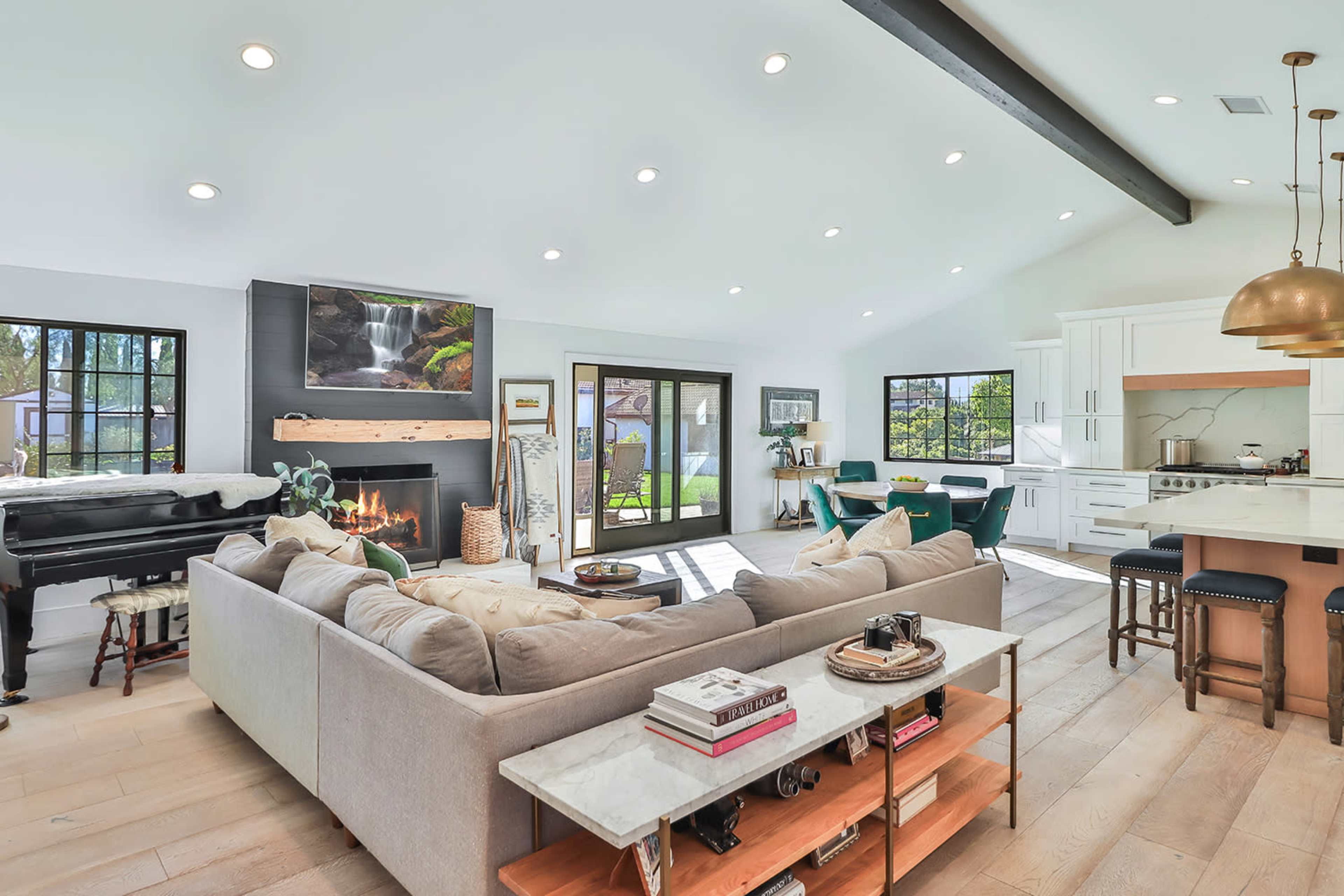 A modern living room with a sectional sofa, a fireplace, a piano, and large windows overlooking a backyard.