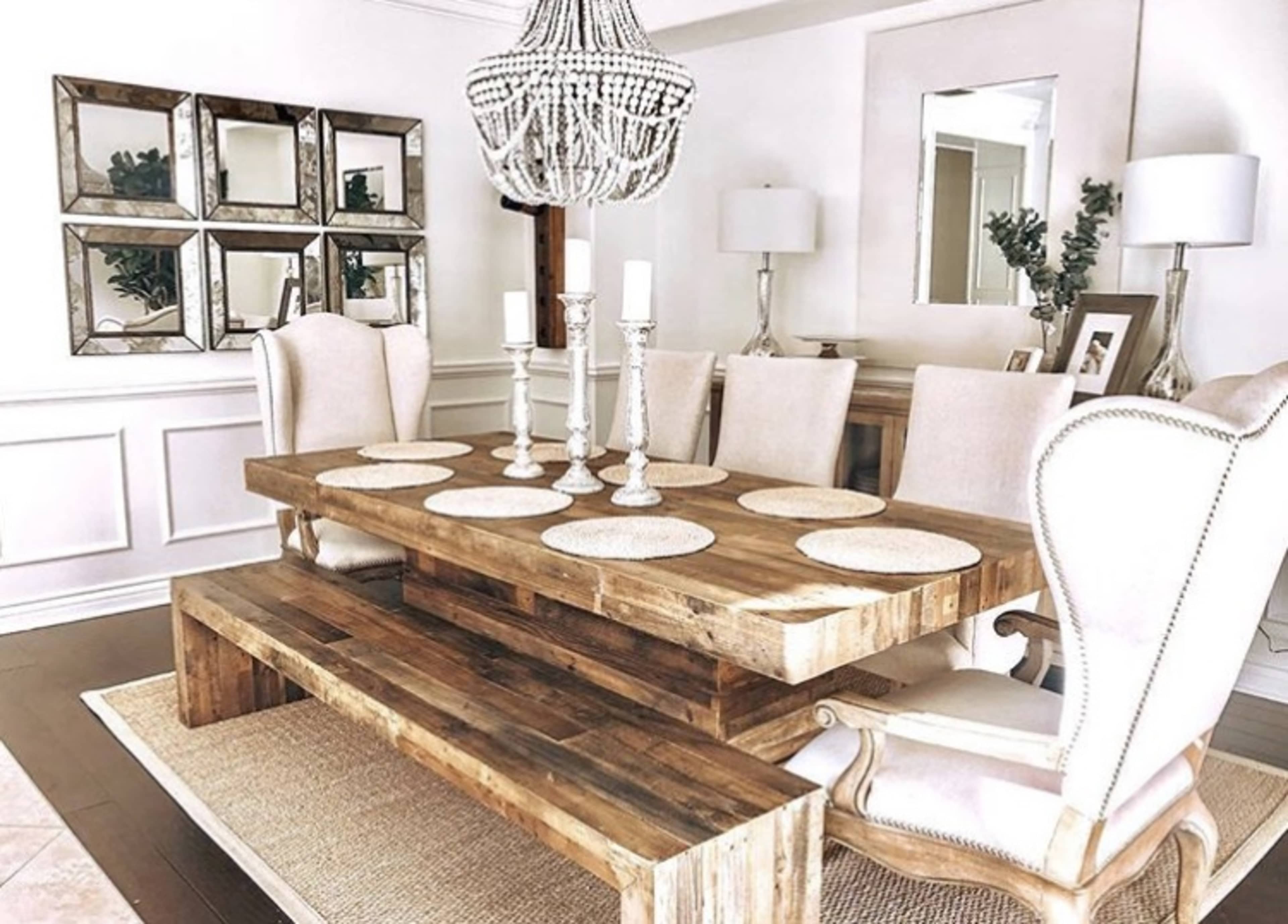 A wooden dining table with upholstered chairs, surrounded by decorative mirrors and candle holders, is set in a bright, elegantly arranged dining room.