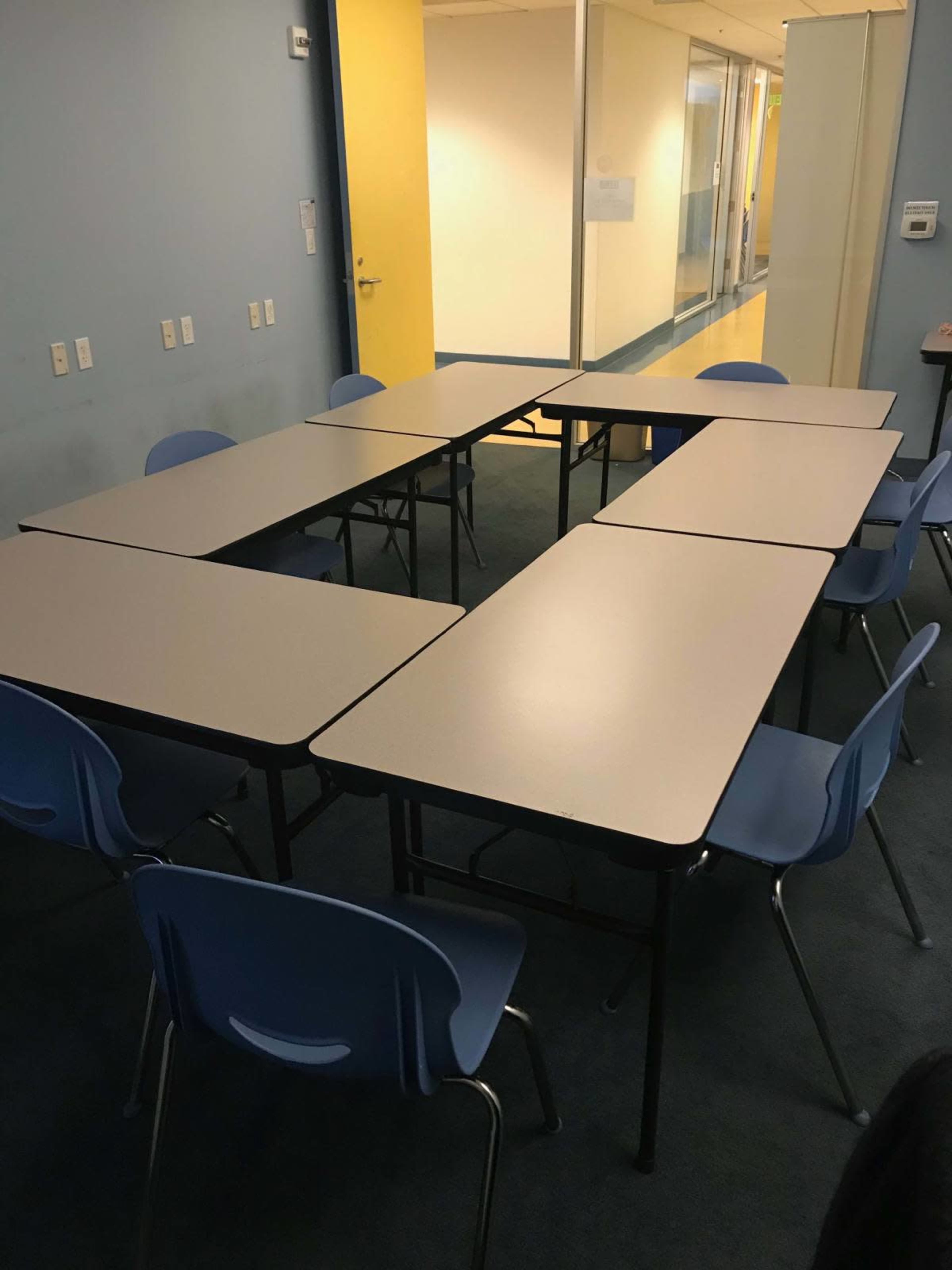 The image shows a rectangular arrangement of tables surrounded by blue chairs in a room with light blue walls and a yellow door.