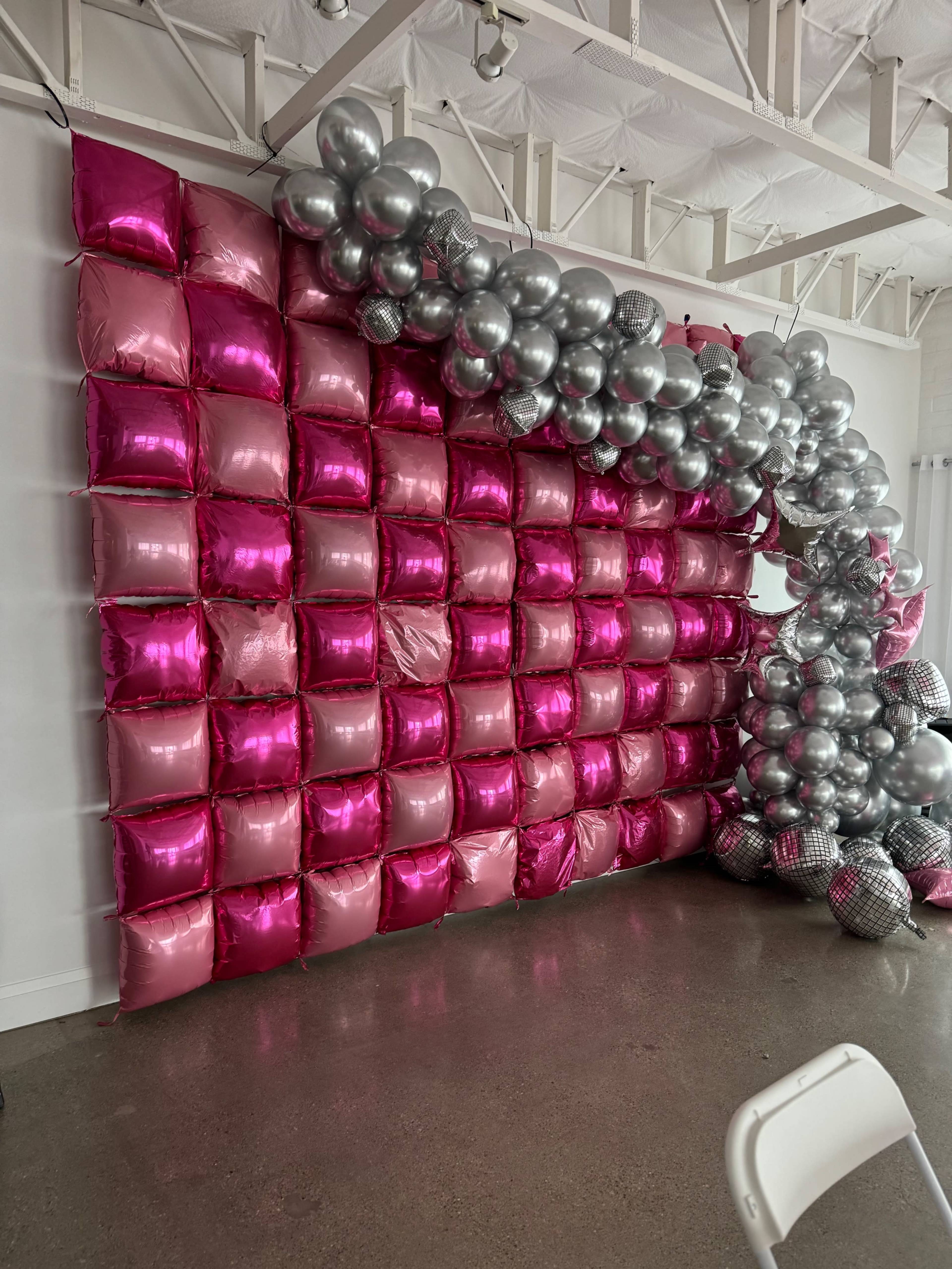 The image shows a decorative wall made of pink and silver balloons arranged in a grid pattern, with a cluster of silver balloons forming an arch at one corner.