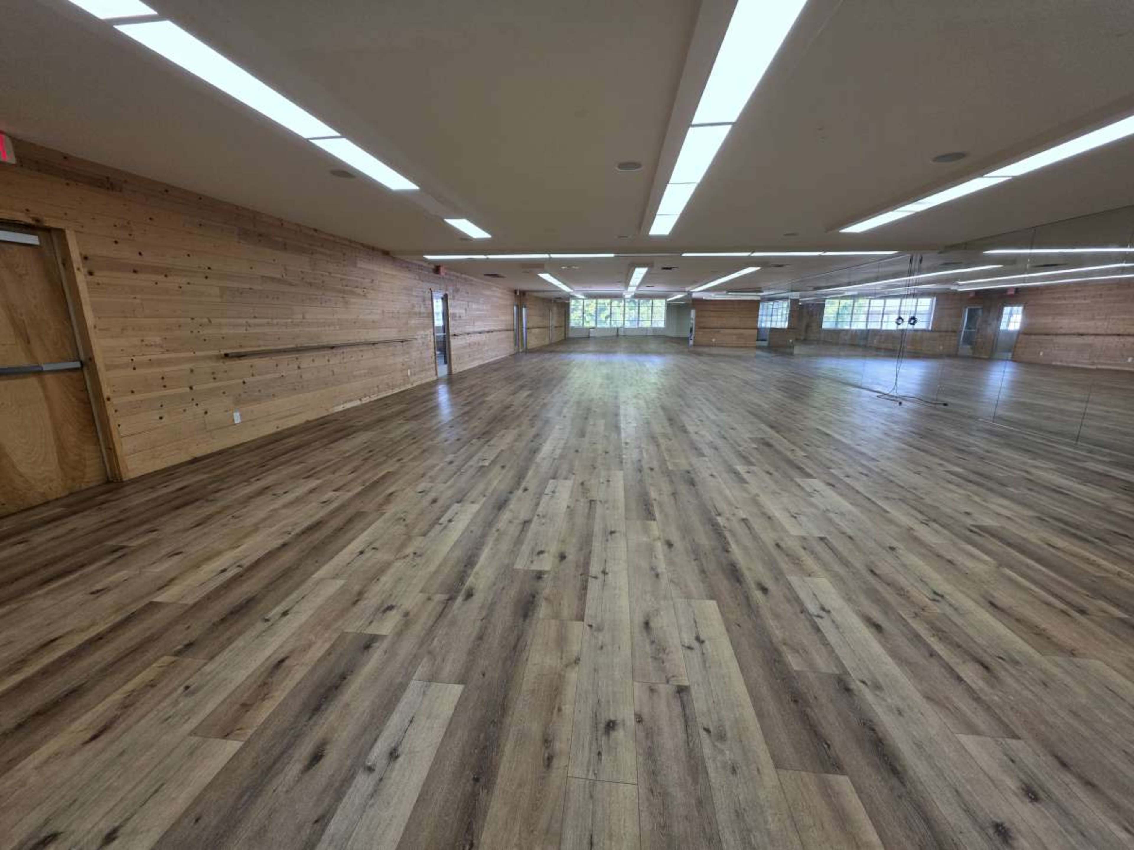 The image shows a spacious wooden-floored room with large mirrors on one wall and natural light streaming in from windows.