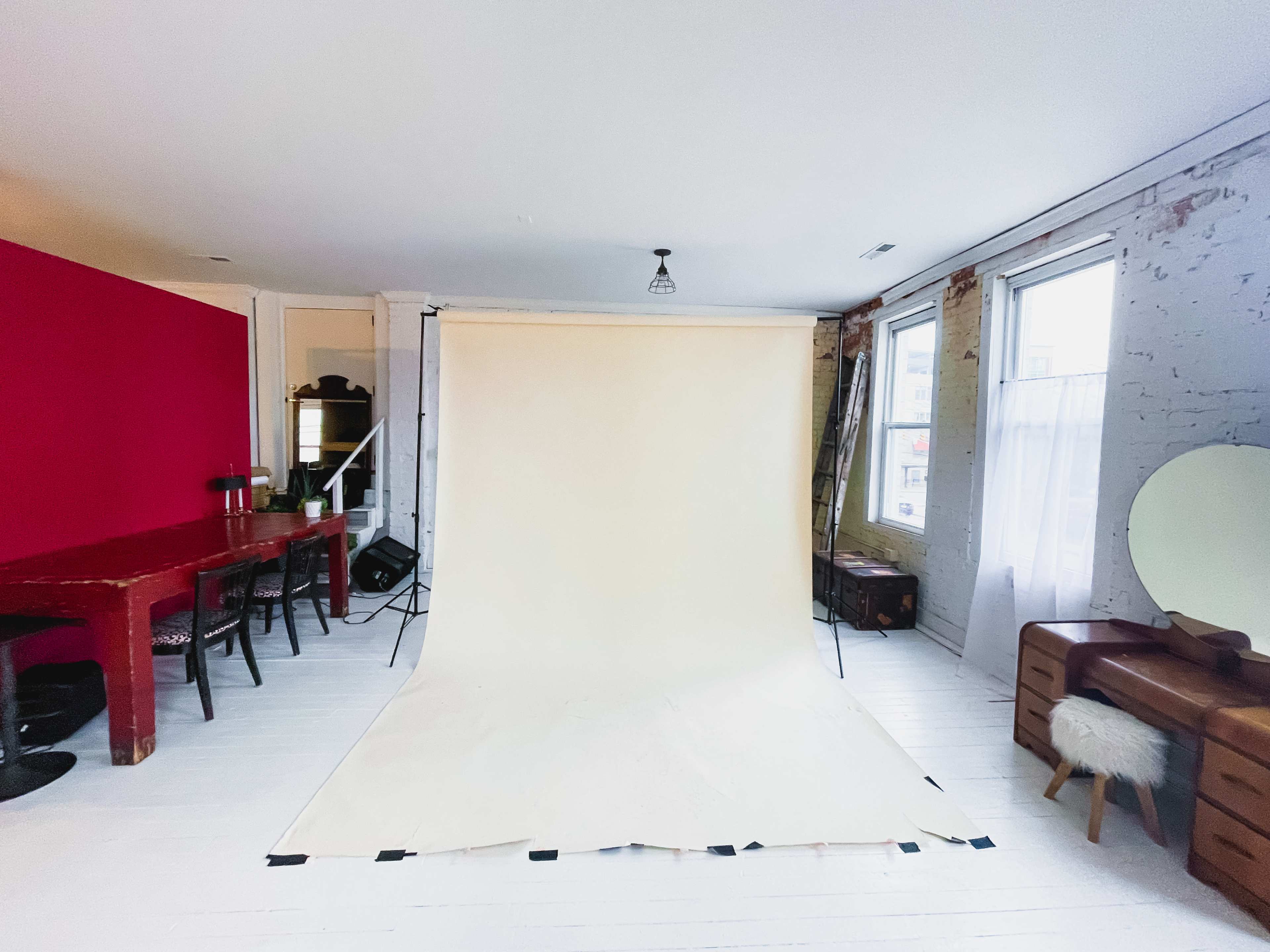 The room features a simple photography setup with a beige backdrop, a red accent wall, and wooden furniture.