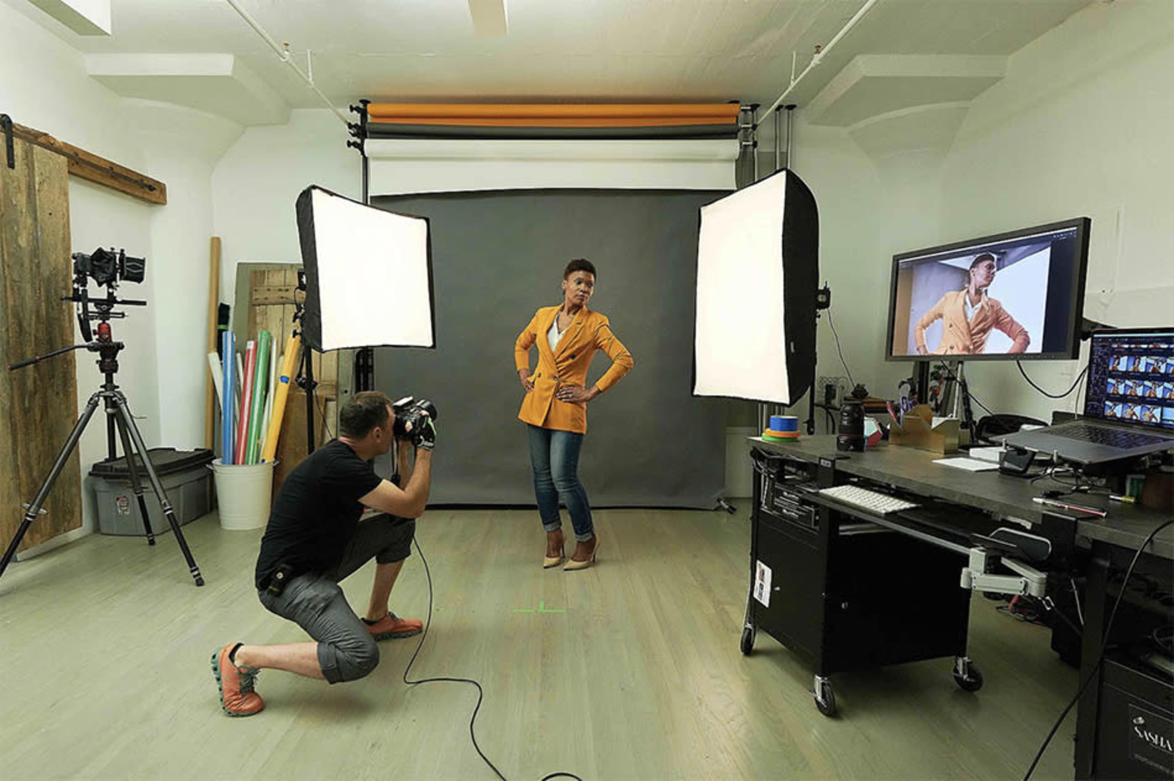A photographer is capturing images of a model in a studio setting with softbox lights and a computer displaying the live feed.