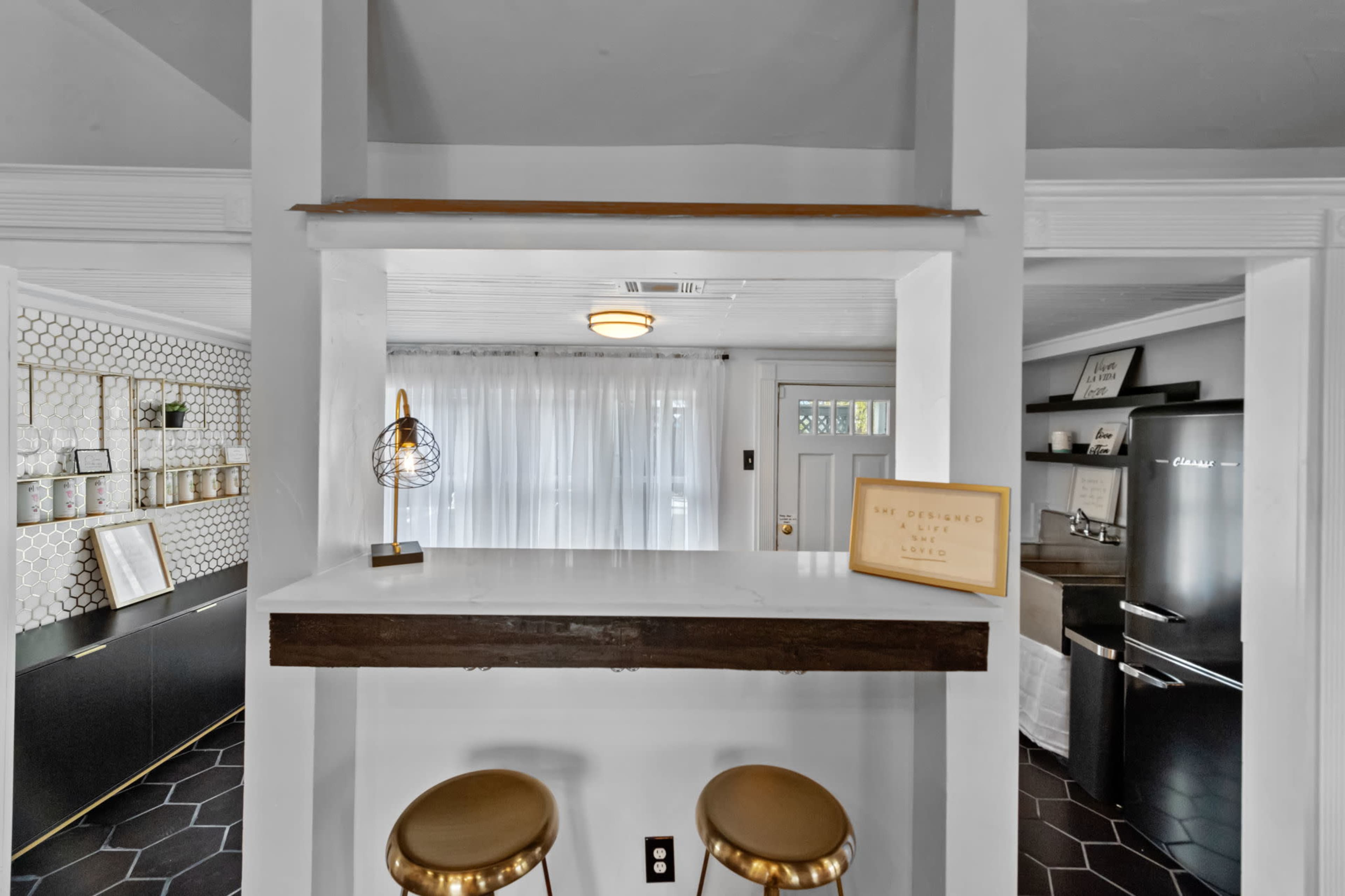 A kitchen space featuring a bar area with two stools, a light fixture, and a view of a living area with white curtains and black cabinetry.