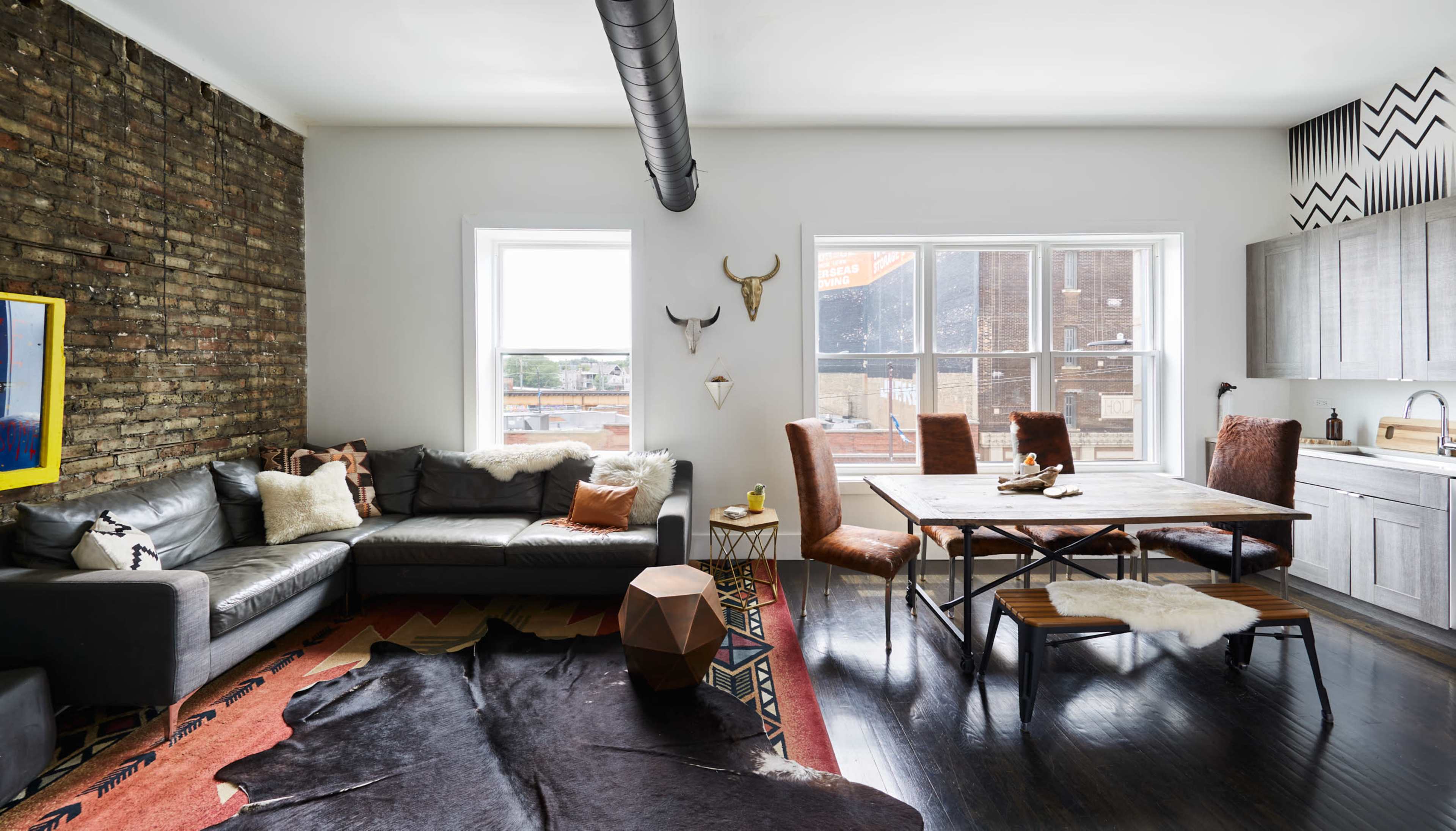 A modern living space features a leather sectional sofa, a wooden dining table surrounded by upholstered chairs, and exposed brick walls with large windows.