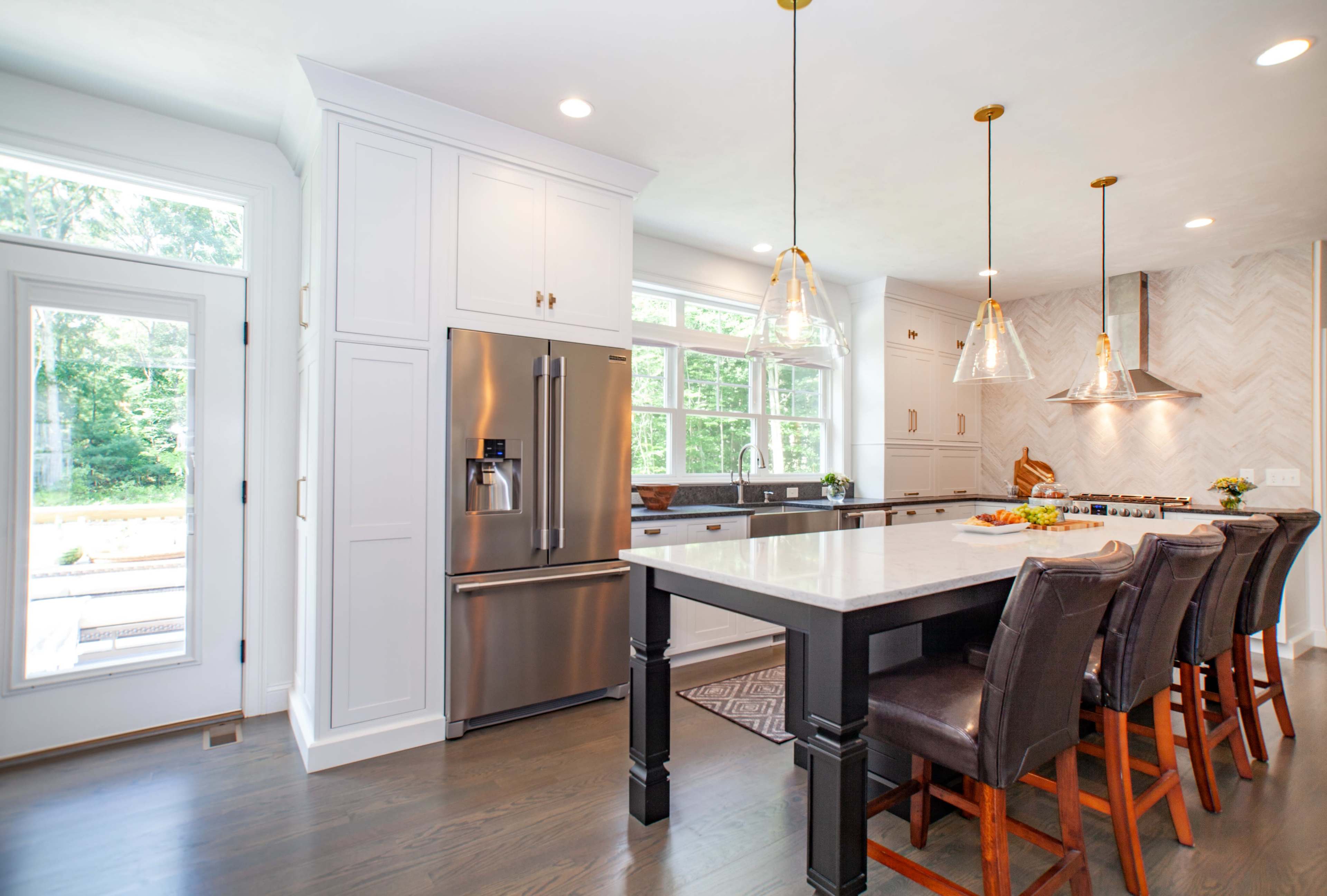 A modern kitchen features a large island with bar seating, stainless steel appliances, white cabinetry, and ample natural light from large windows.