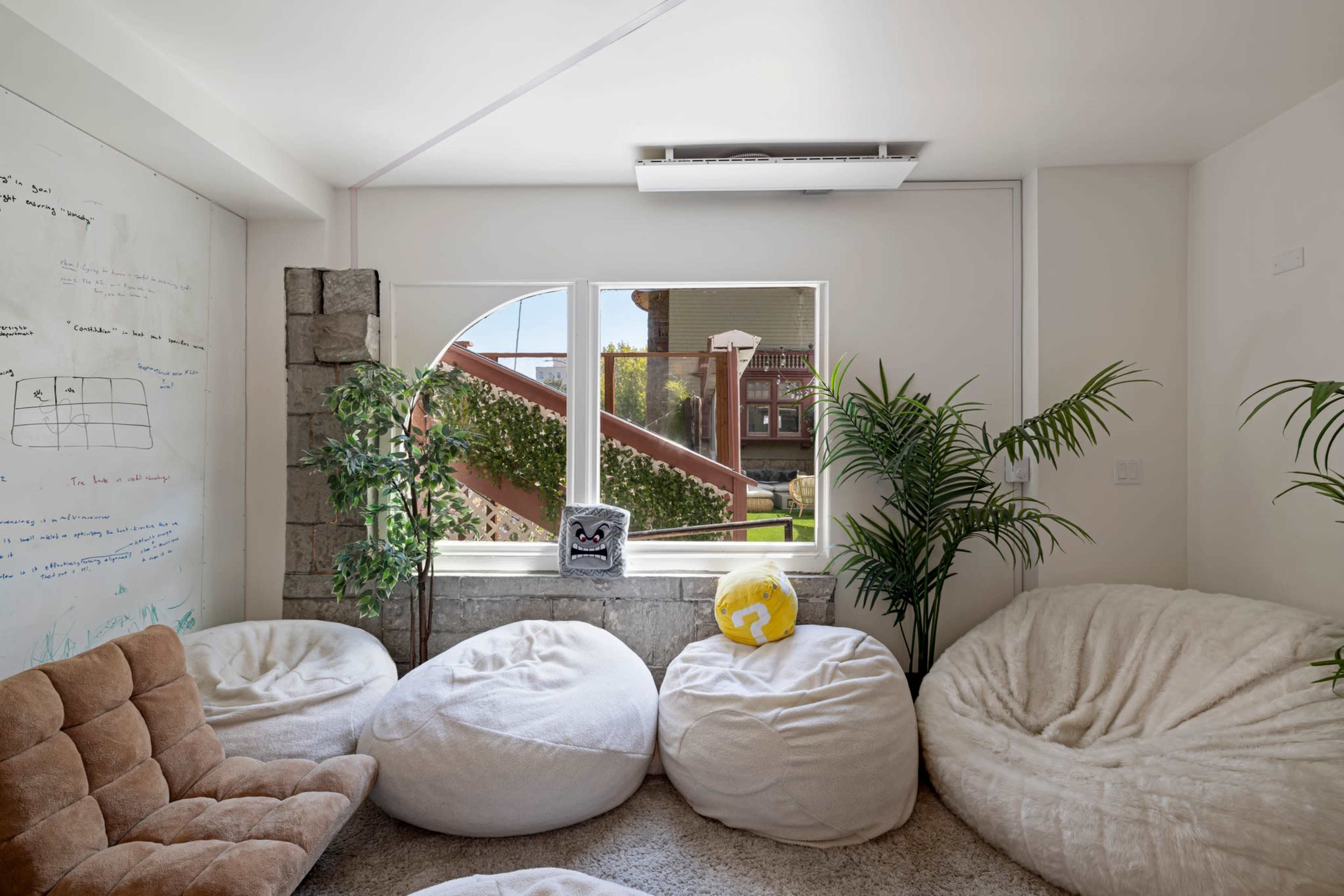 The room features several large bean bags arranged around a window, with plants beside a concrete wall and whiteboard covered in text.