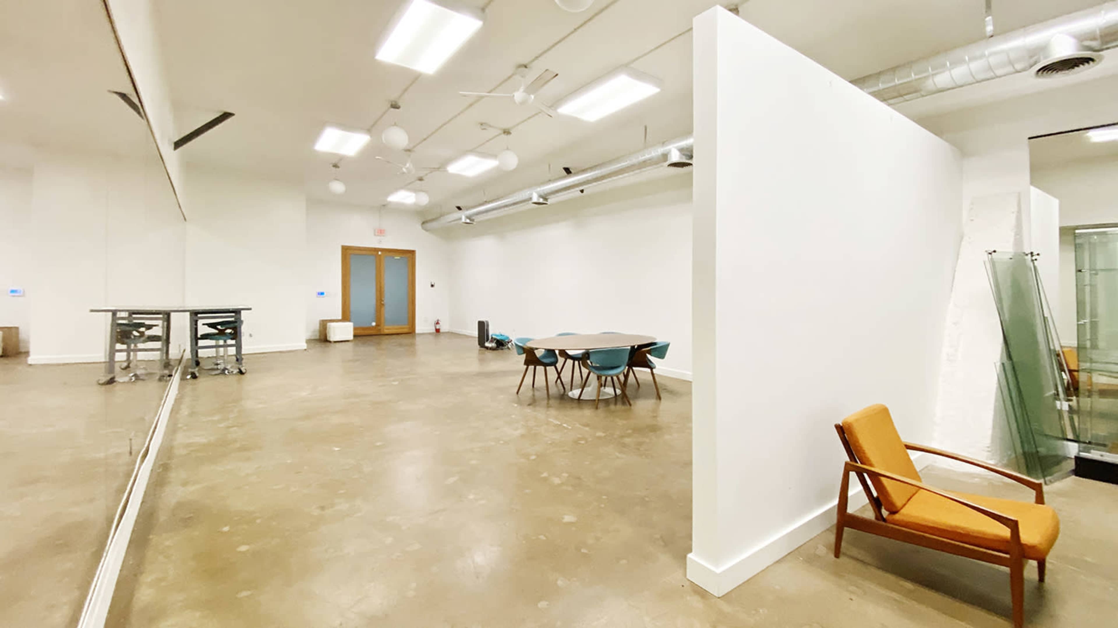 The image shows a spacious, minimalist room with a large table surrounded by chairs, a brown armchair in the corner, and large mirrors along one wall.