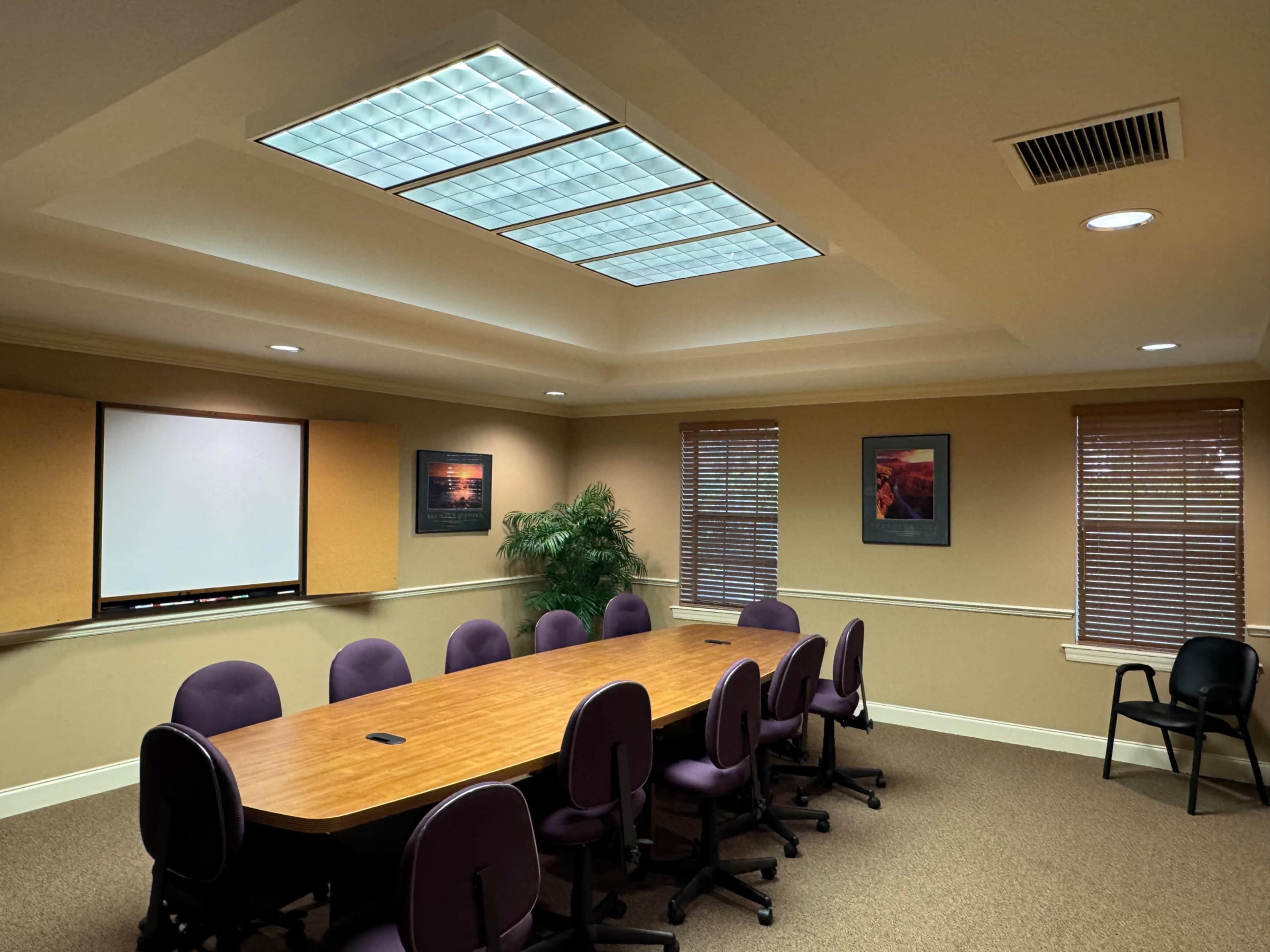 A conference room with a long wooden table surrounded by purple chairs, a whiteboard, and two framed pictures on the walls.