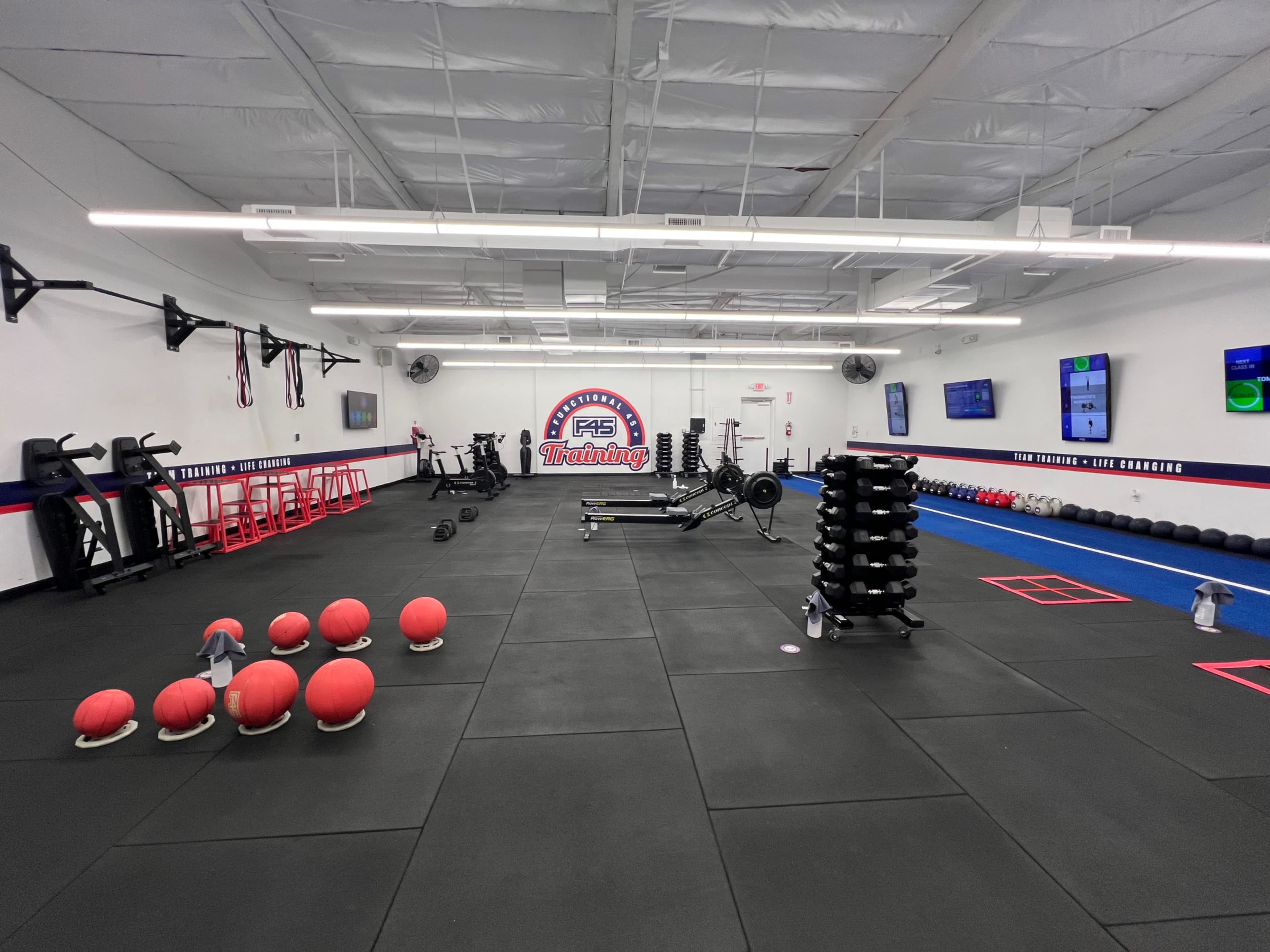 The image shows an empty gym space with workout equipment including weights, benches, and exercise balls arranged on a rubber floor.