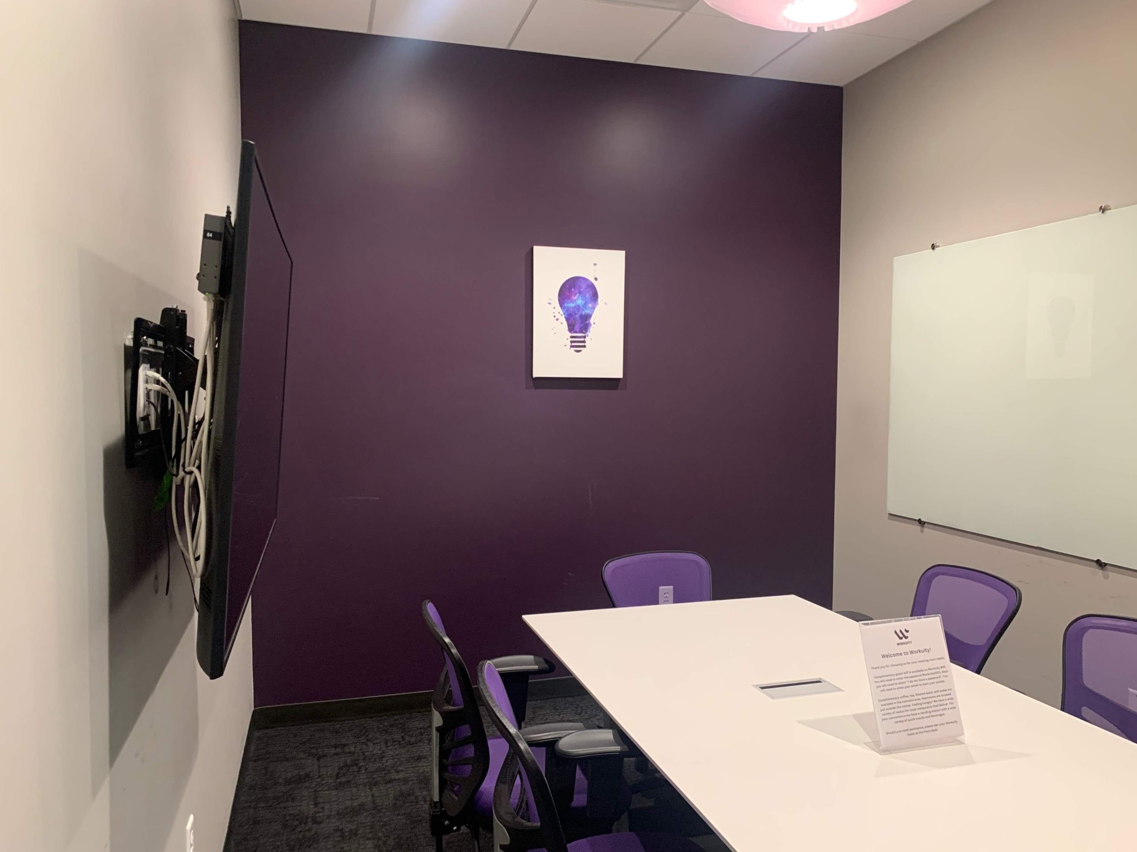A small conference room features a white table, four purple chairs, a wall-mounted TV, and a light bulb-themed artwork on a deep purple wall.